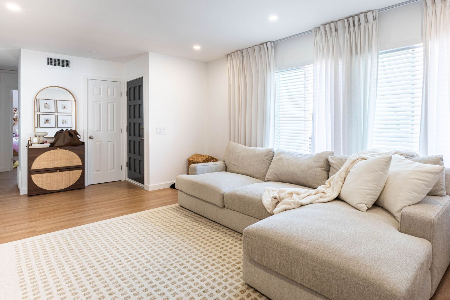 Bright living room with beige sectional sofa, hardwood floors, patterned rug, and large windows with sheer curtains.