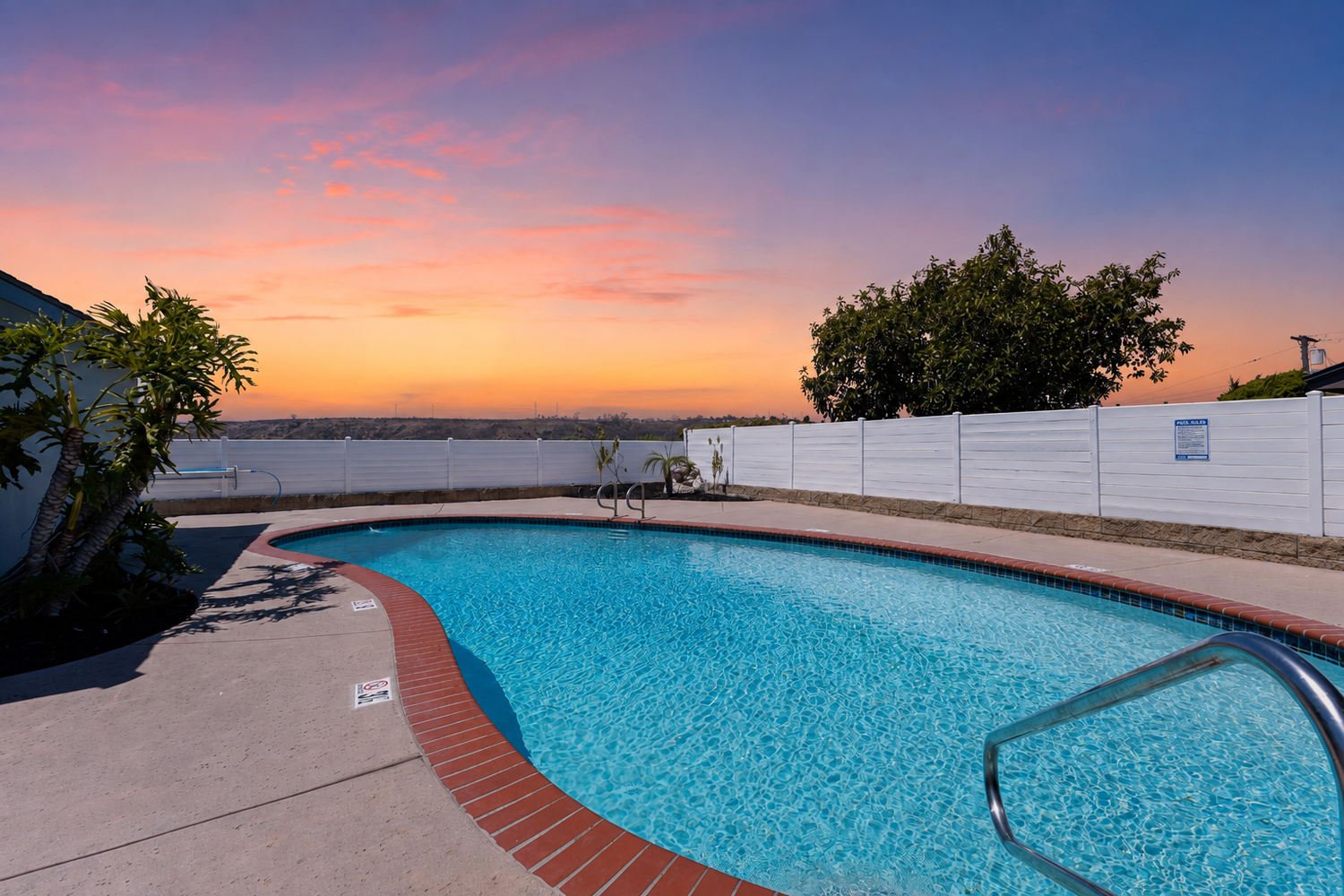 Stunning outdoor pool with crystal-clear water reflecting a vibrant sunset sky, surrounded by a white fence and lush trees.