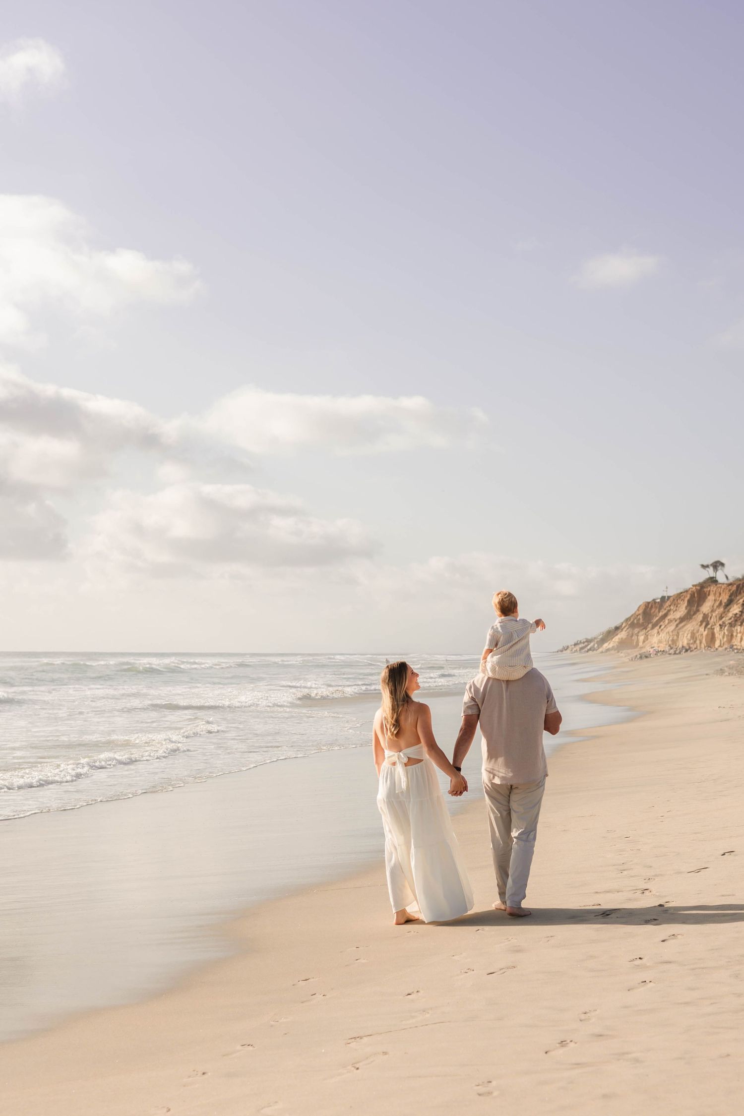 A family walks along a sandy beach, with a child sitting on dad's shoulders, holding hands at sunset.