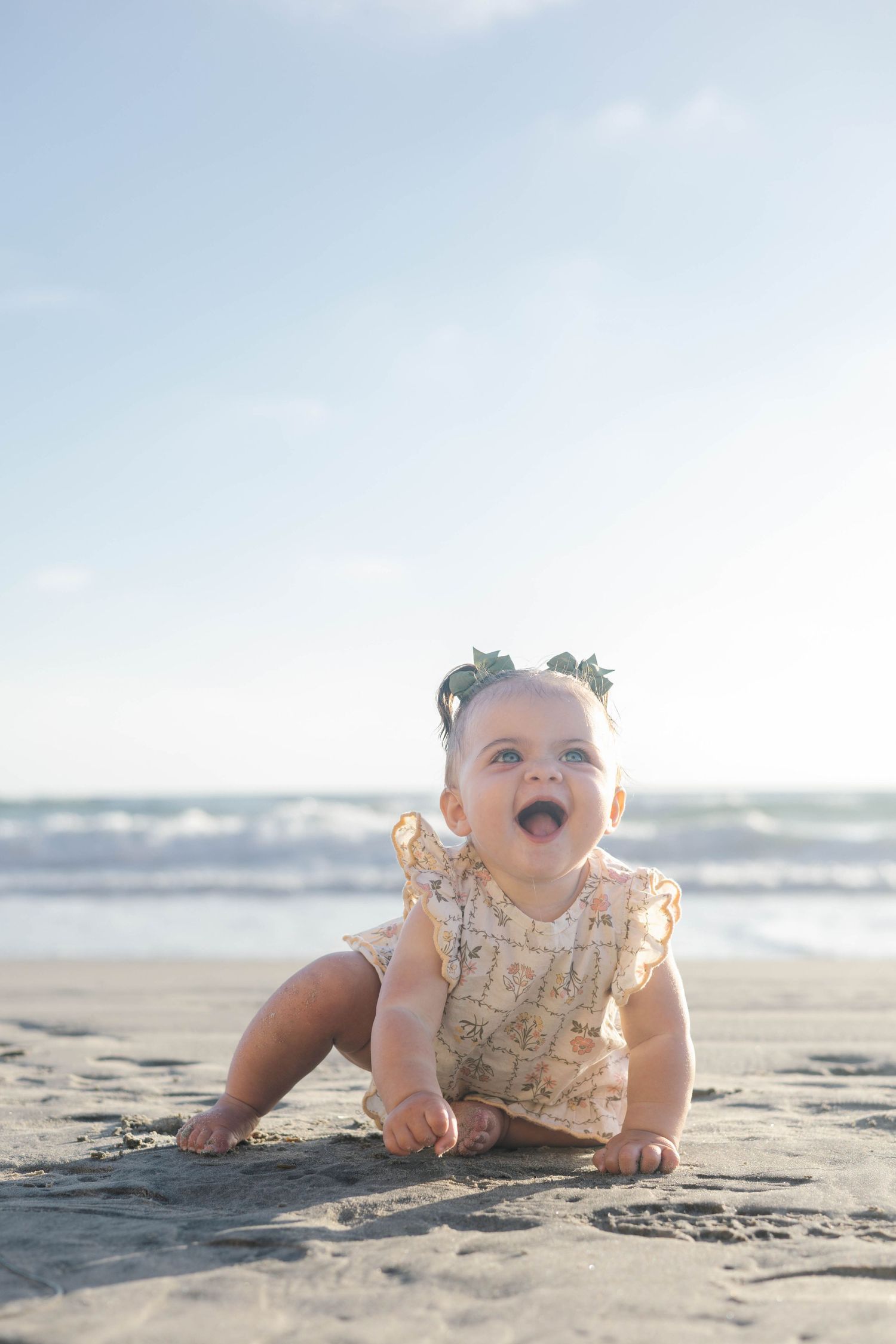 Happy baby girl sitting on sandy beach wearing floral romper and flower headband with ocean waves behind her.