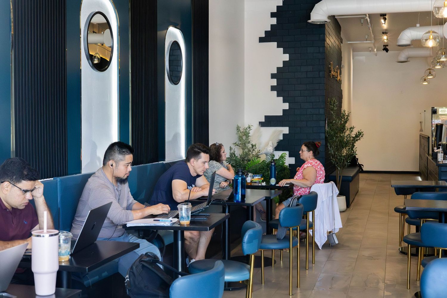 Modern cafe interior with blue velvet booths and chairs, patrons working on laptops in a stylish contemporary setting.