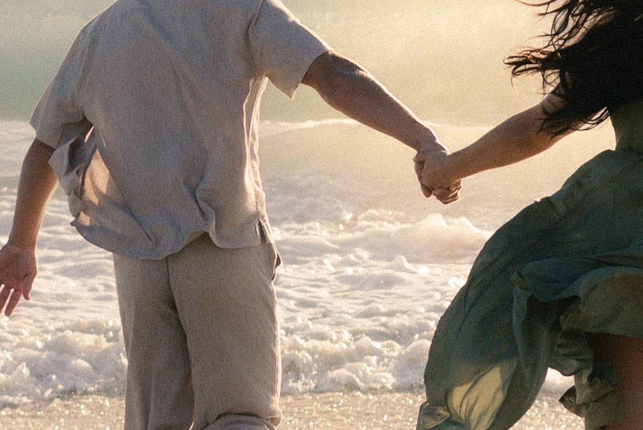 Couple holding hands running toward ocean waves at sunset, womans green dress flowing in the breeze.