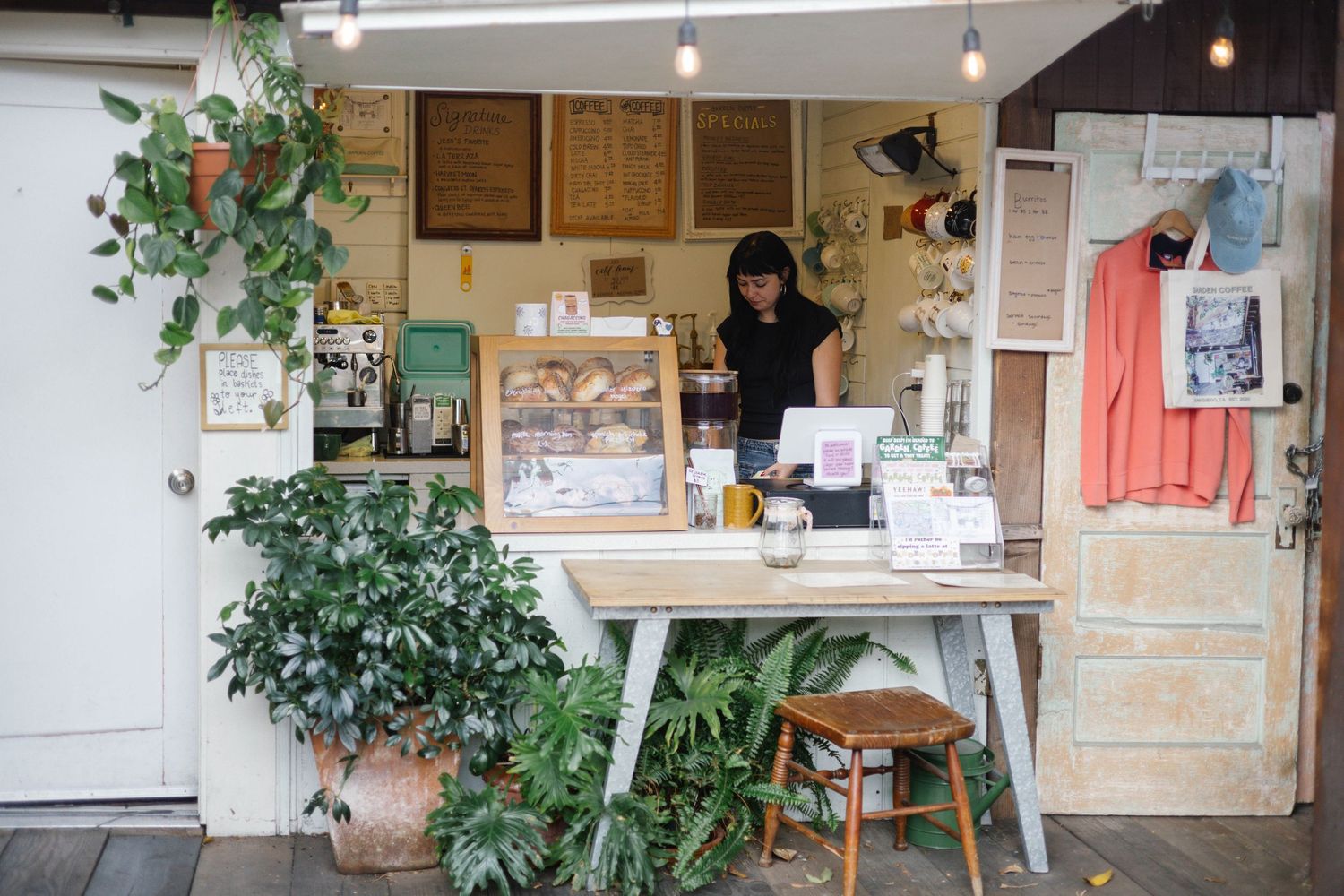 A woman stands behind a rustic cafe counter surrounded by lush green plants and warm Edison bulb lighting.