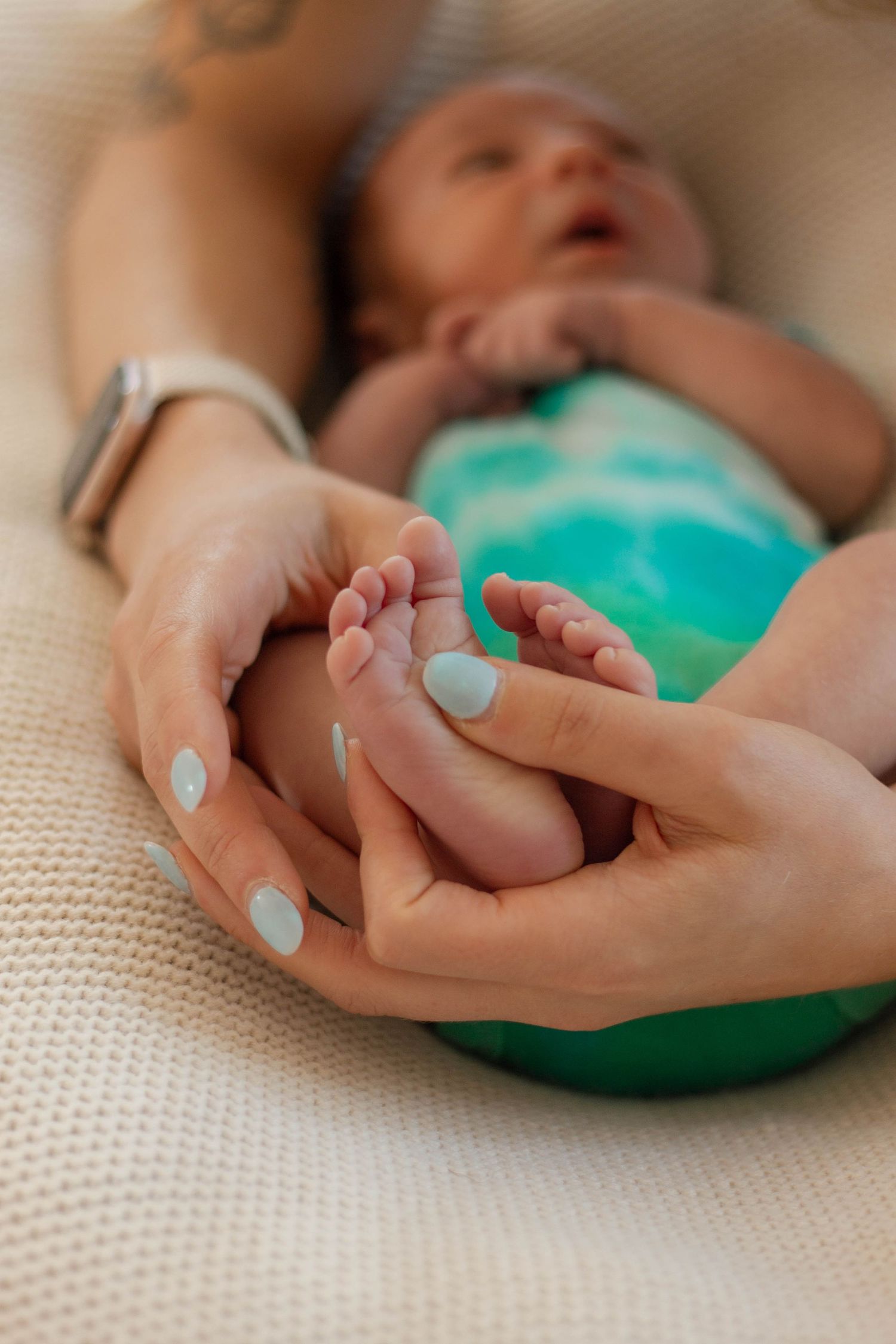 Close-up of mother's hands gently holding newborn baby's tiny feet in teal outfit.