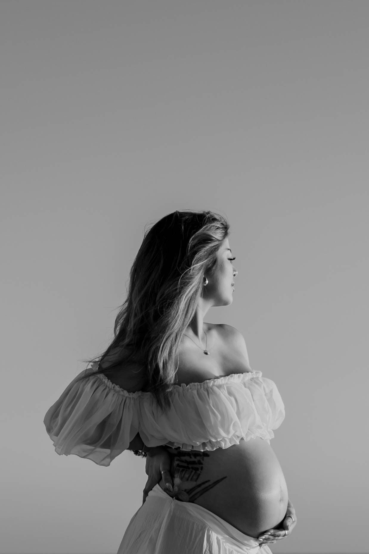 Black and white portrait of pregnant woman in off-shoulder dress holding baby bump.