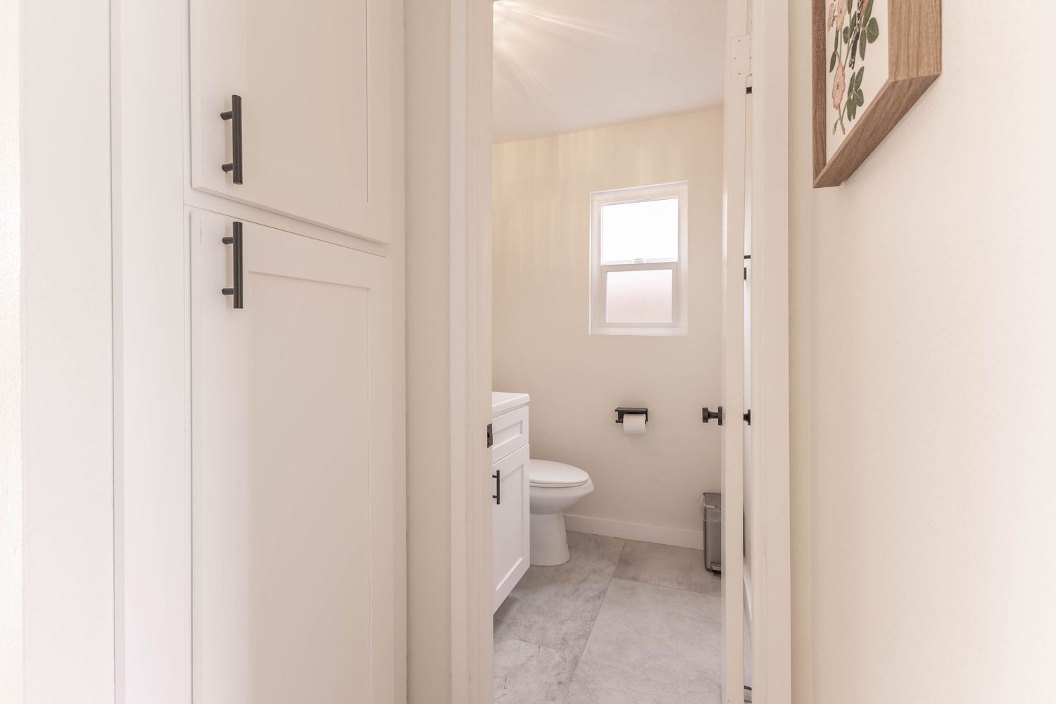 Bright white hallway leading to a clean bathroom with toilet, marble tile floors, and a small window.