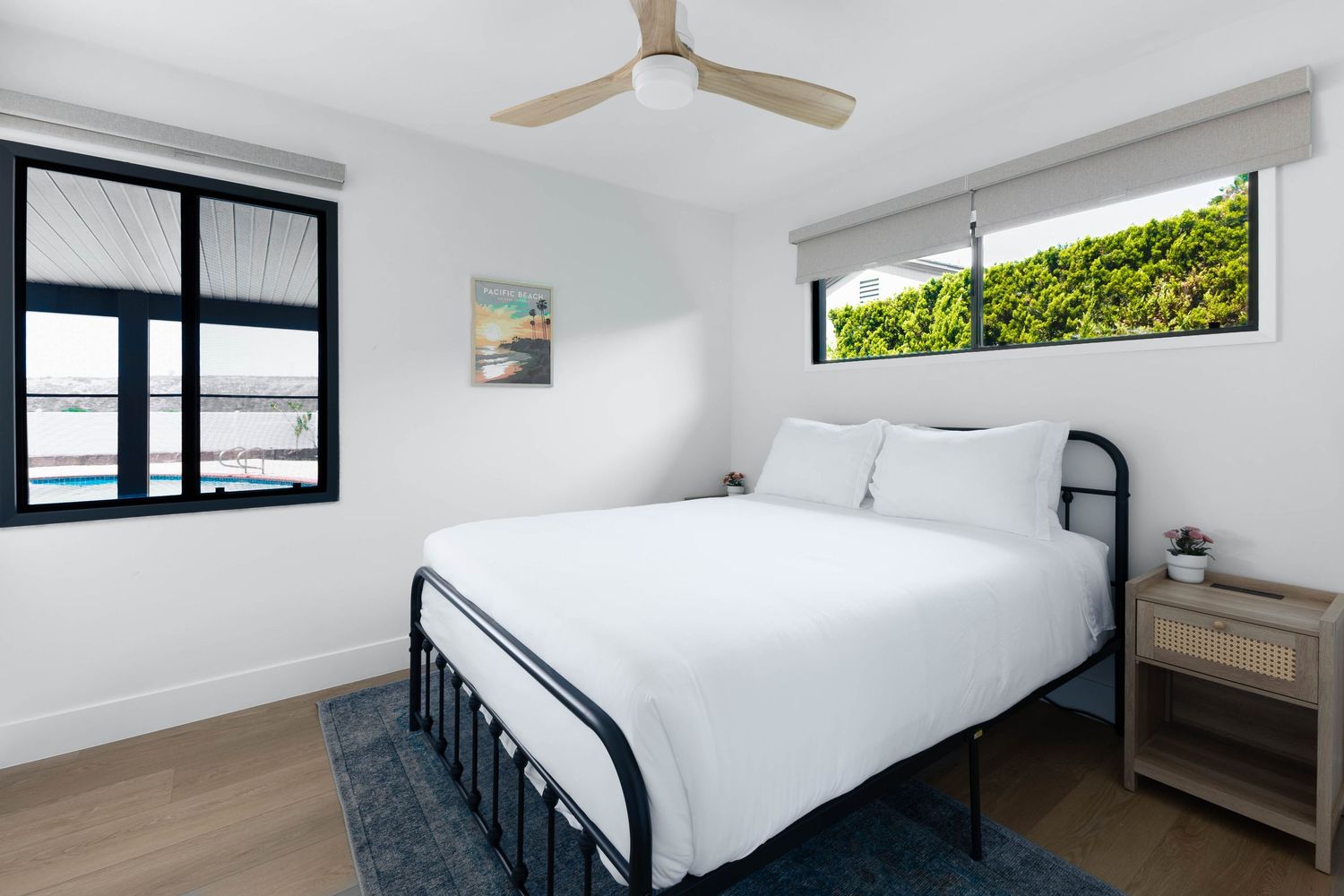 Modern bedroom with black metal bed frame, white bedding, hardwood floors, ceiling fan, and bright windows with garden views.
