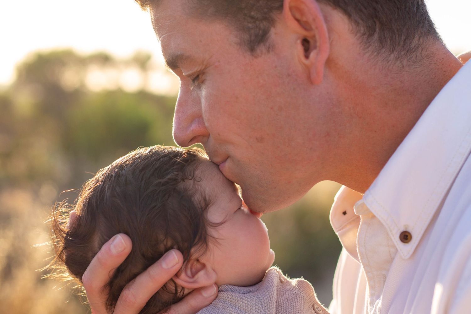 Tender moment of father gently kissing newborn babys head in warm golden outdoor light.