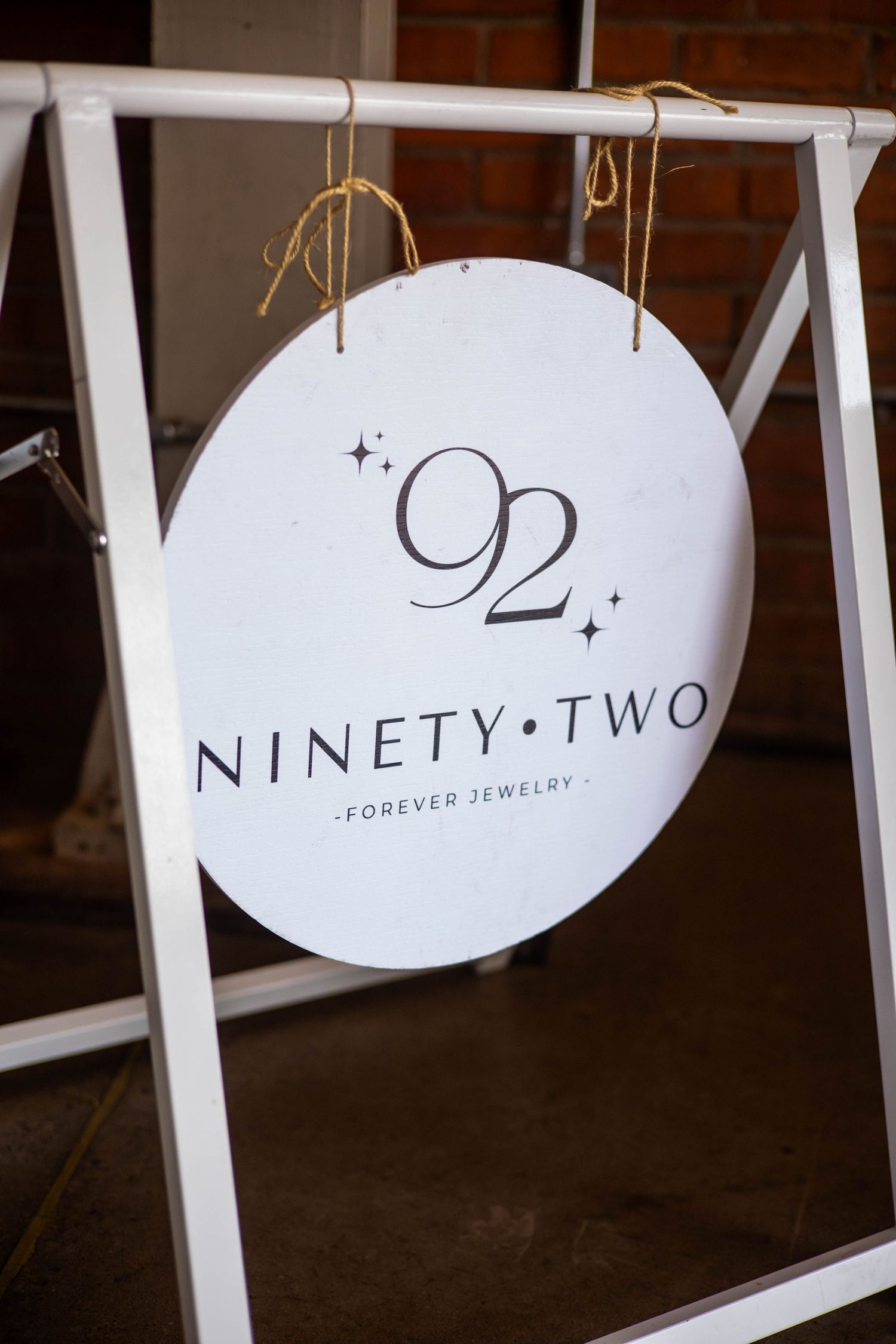 Round white sign for 92 Ninety-Two Forever Jewelry hanging from a white frame display stand.