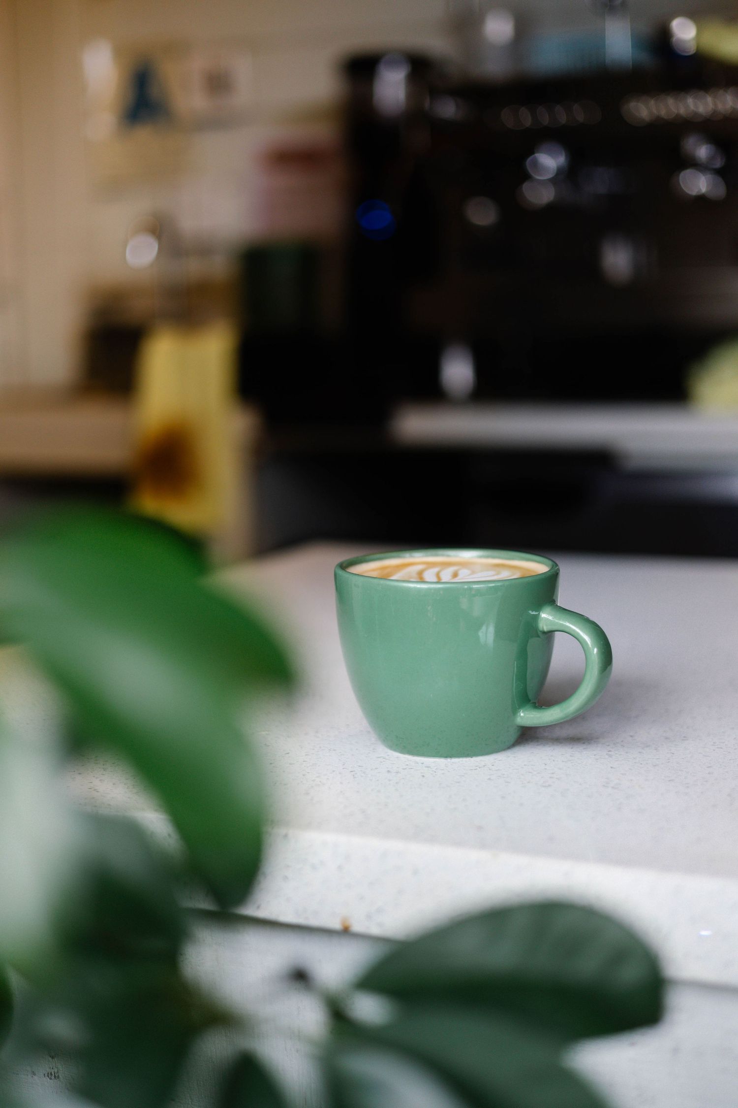 A green ceramic coffee cup with latte art sits on a white counter in a modern cafe with blurred greenery.