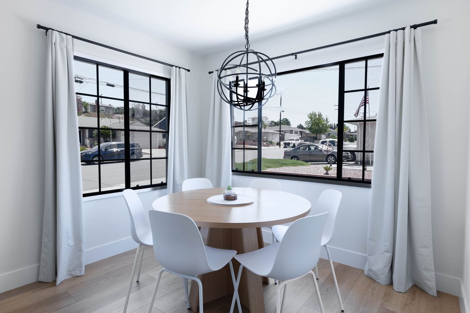 Modern dining nook with round wood table, white chairs, black-framed windows, and an orb chandelier on light hardwood floors.