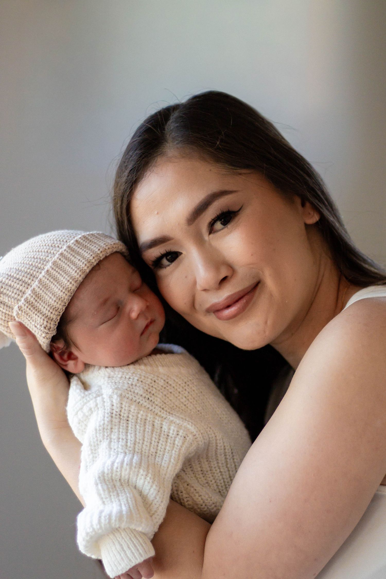 Young Asian mother smiling and holding sleeping newborn baby dressed in knit outfit and beanie hat.