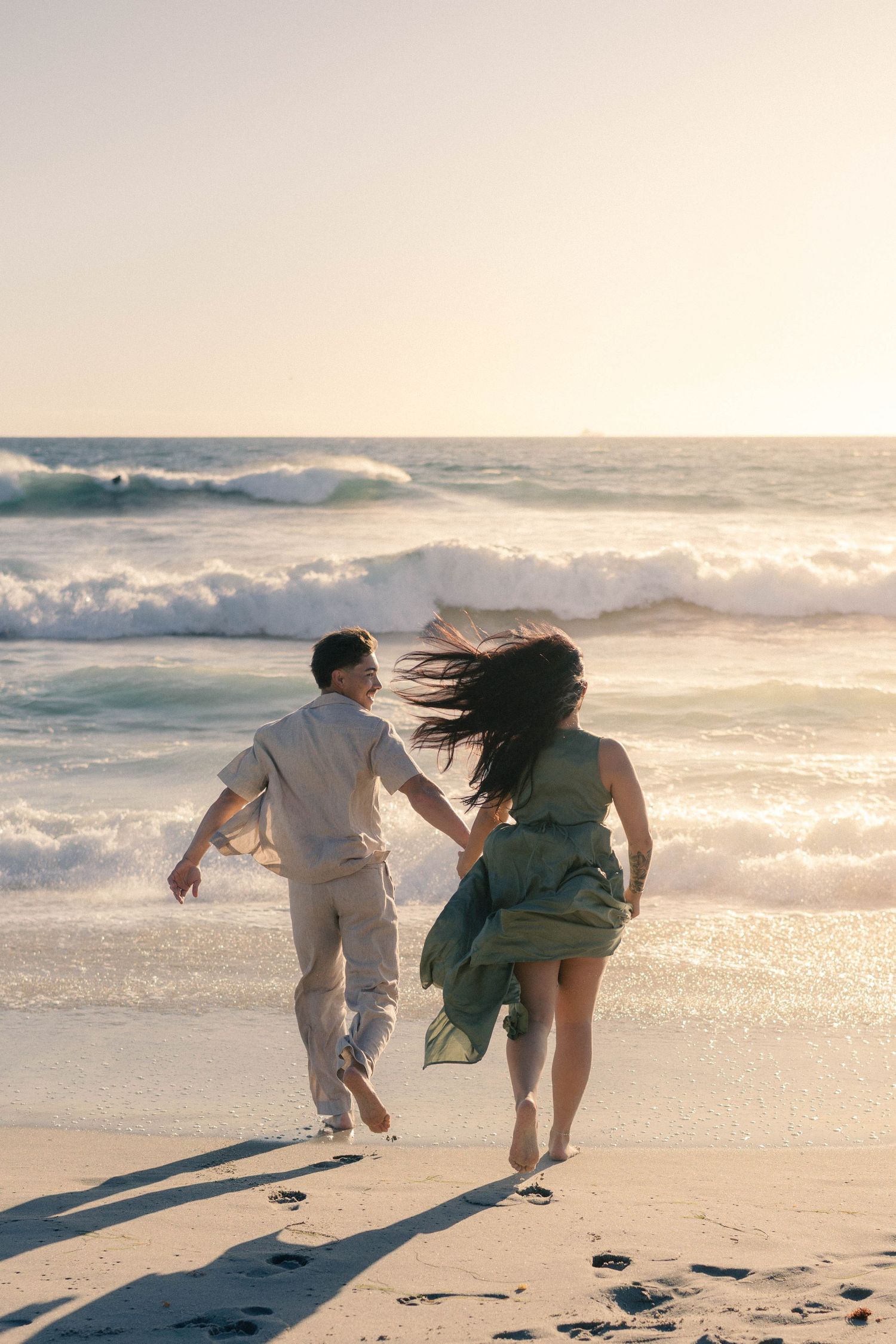 Couple running hand-in-hand toward crashing ocean waves at sunset on a sandy beach.