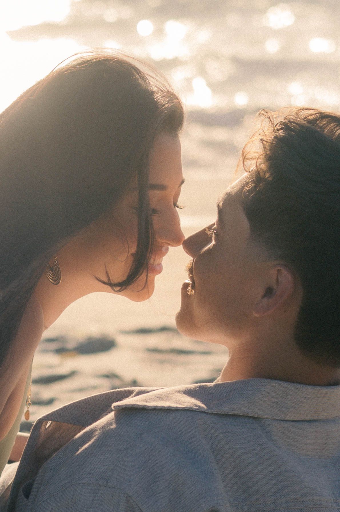 Close-up of couple almost kissing at golden sunset by the ocean, bathed in warm glowing light.