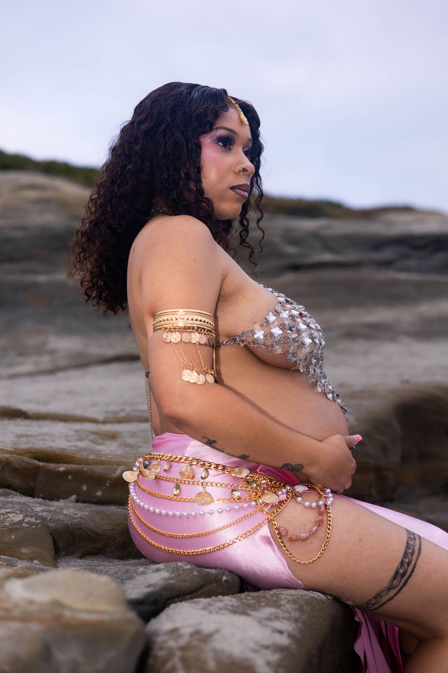 Pregnant woman in sequined top and pink skirt with gold chain accessories sits on rocks outdoors.