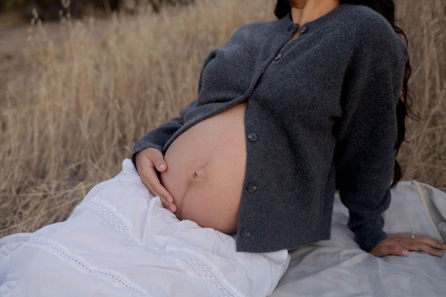 Pregnant woman in gray cardigan sitting outdoors in a field, cradling her bare baby bump with both hands.