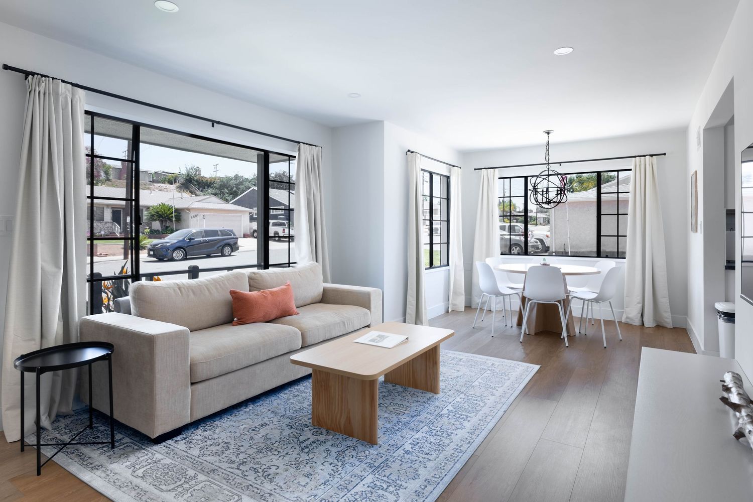 Modern open-plan living room with beige sofa, wood coffee table, blue rug, and dining area with chandelier and large windows.