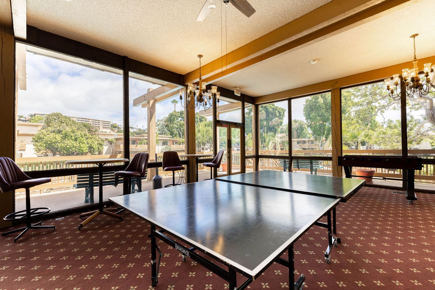 Bright sunroom with ping pong table, chandelier lighting, large windows, and patio views on patterned carpet flooring.