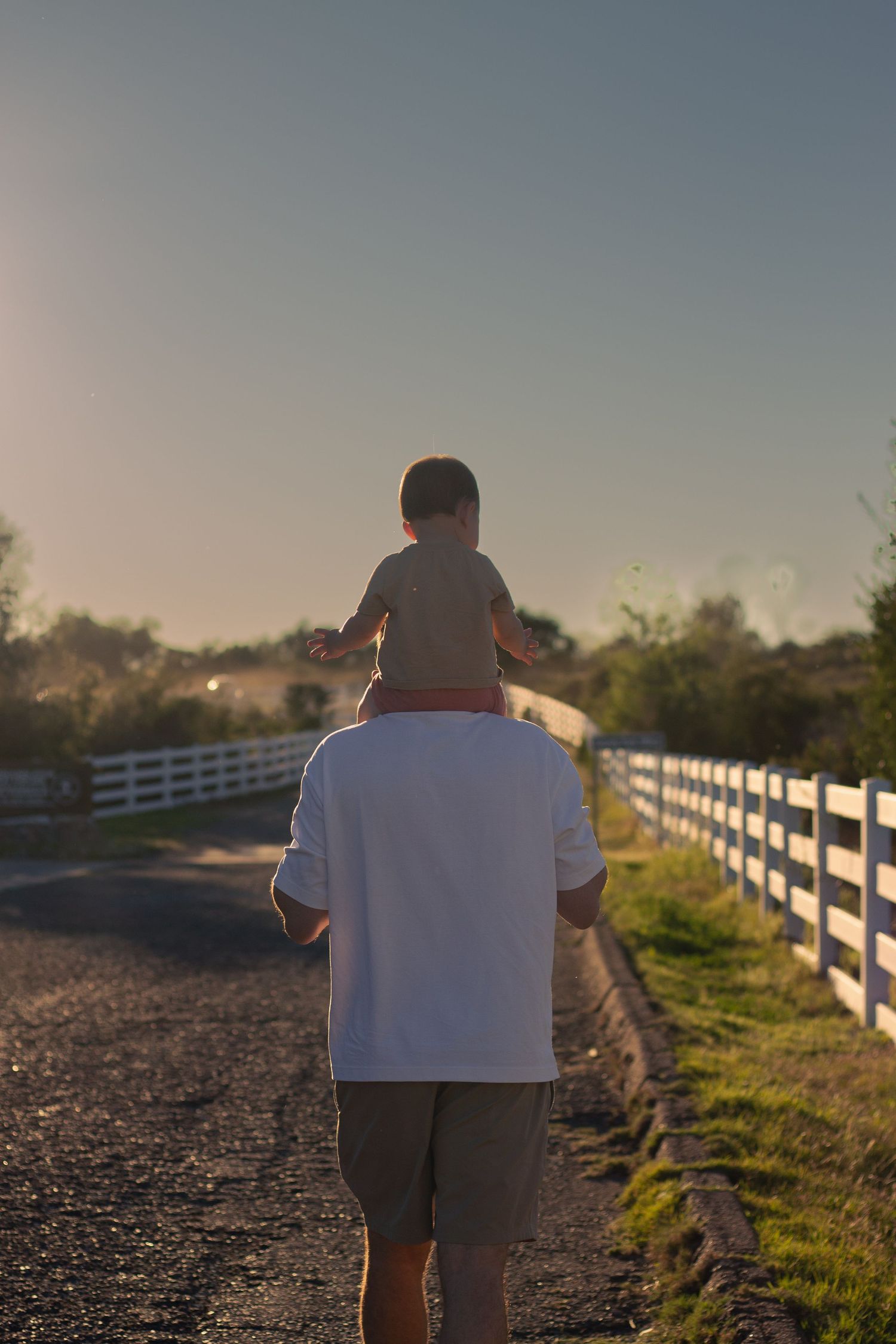 A parent carries a young child on their shoulders while walking along a sunlit fenced road.