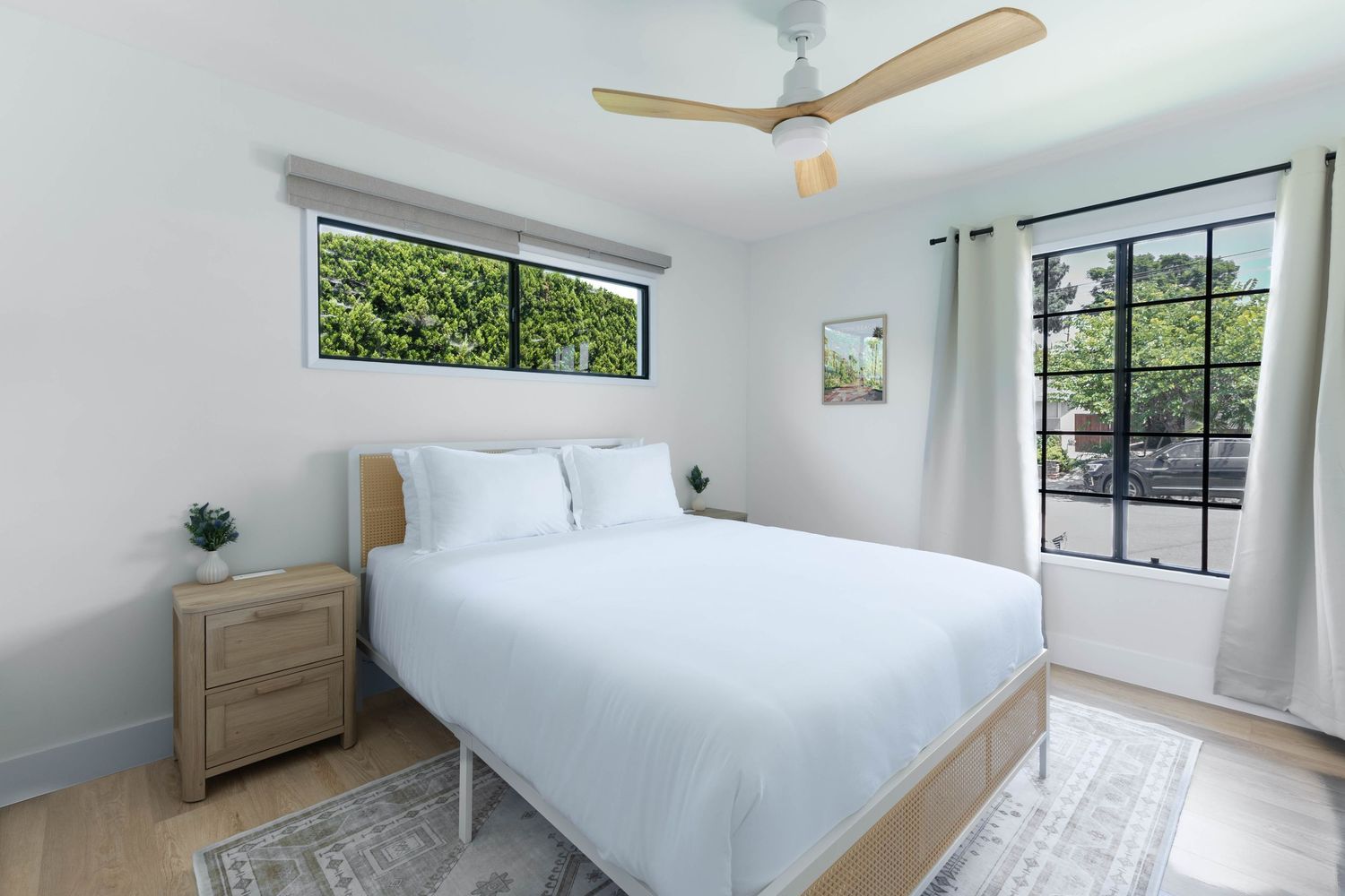 Modern bedroom with white bedding, wood nightstand, ceiling fan, and large windows with green views.