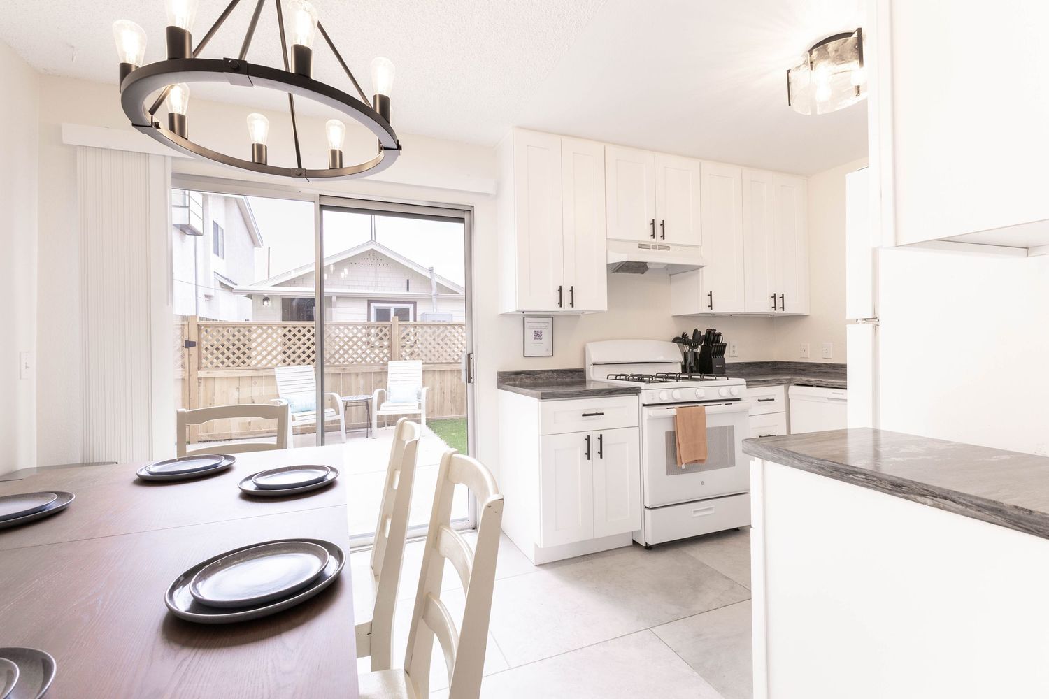 White kitchen with dark countertops and pendant light fixture, open to dining area with sliding door to backyard.