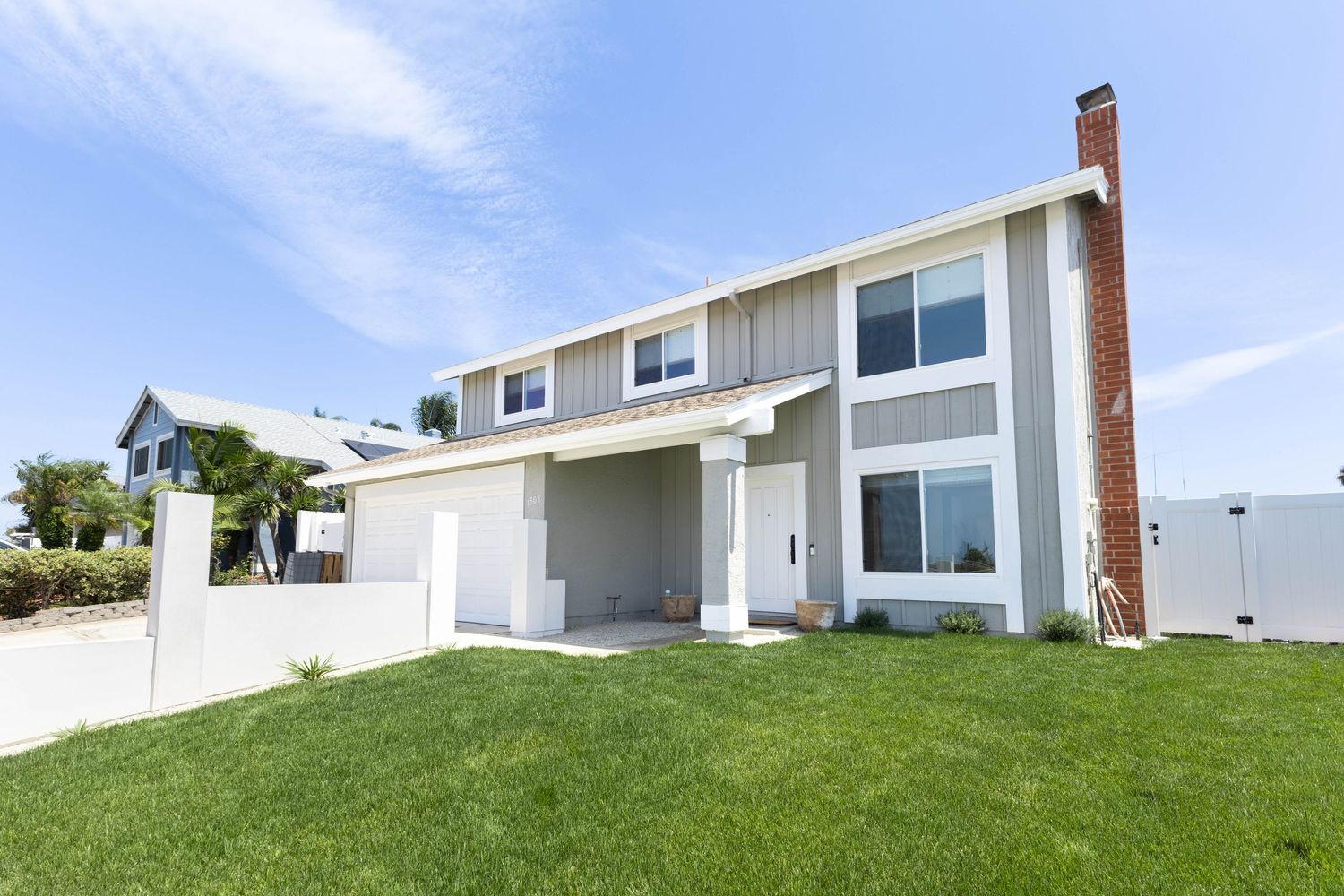 Two-story suburban home with white and gray siding, brick chimney, attached garage, and lush green lawn under blue sky.