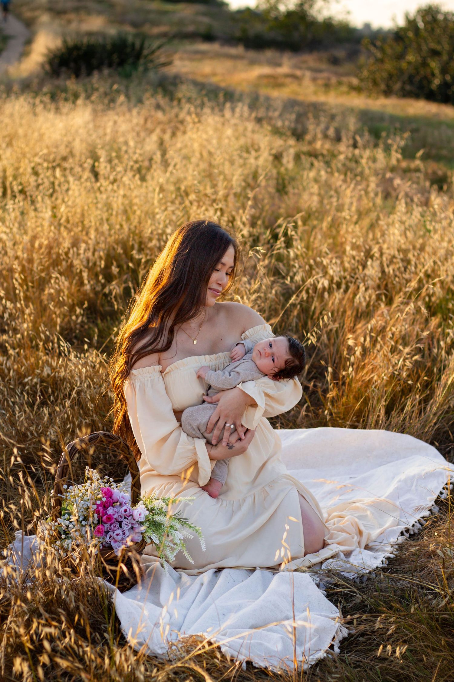 Mother in cream off-shoulder dress sits in golden field cradling sleeping newborn at sunset.