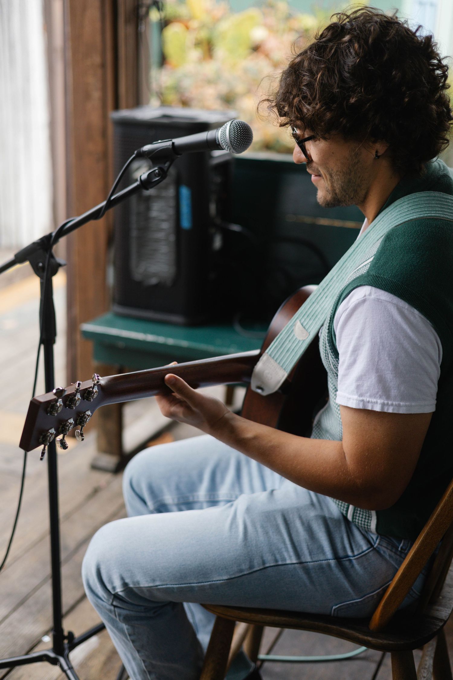 Young man with curly hair playing acoustic guitar while singing into a microphone outdoors.