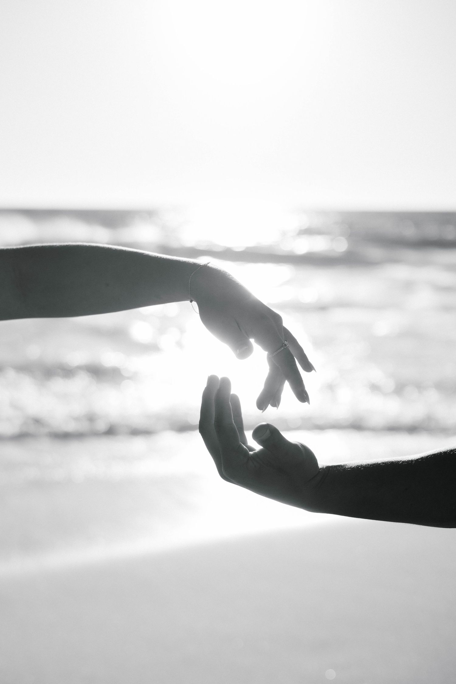 Black and white artistic shot of two hands reaching toward each other with ocean and sunlight behind.