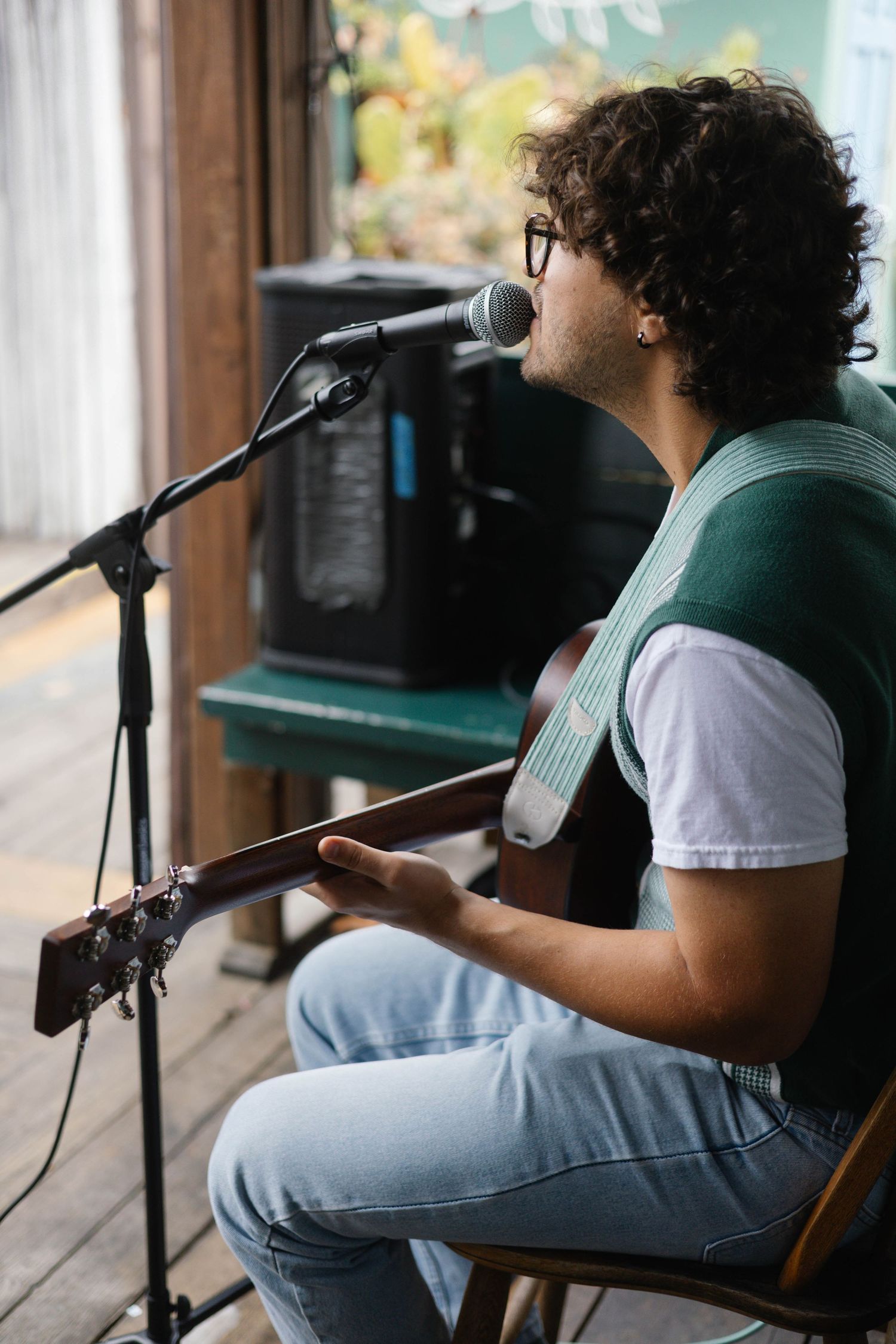 Young curly-haired musician playing acoustic guitar and singing into microphone at outdoor cafe.