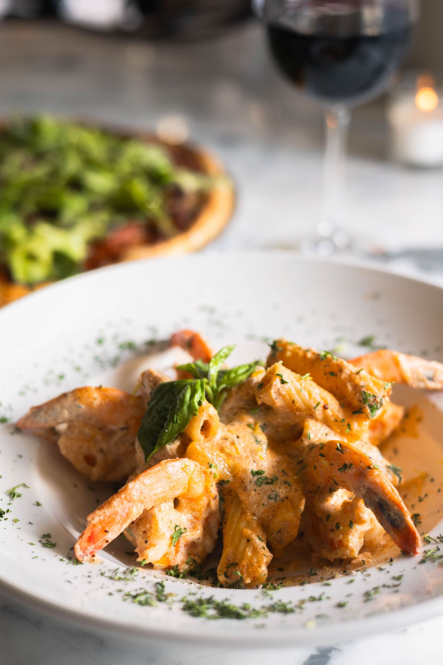 Creamy pasta with shrimp and broccoli garnished with fresh herbs in a white bowl, with red wine and pizza in background.