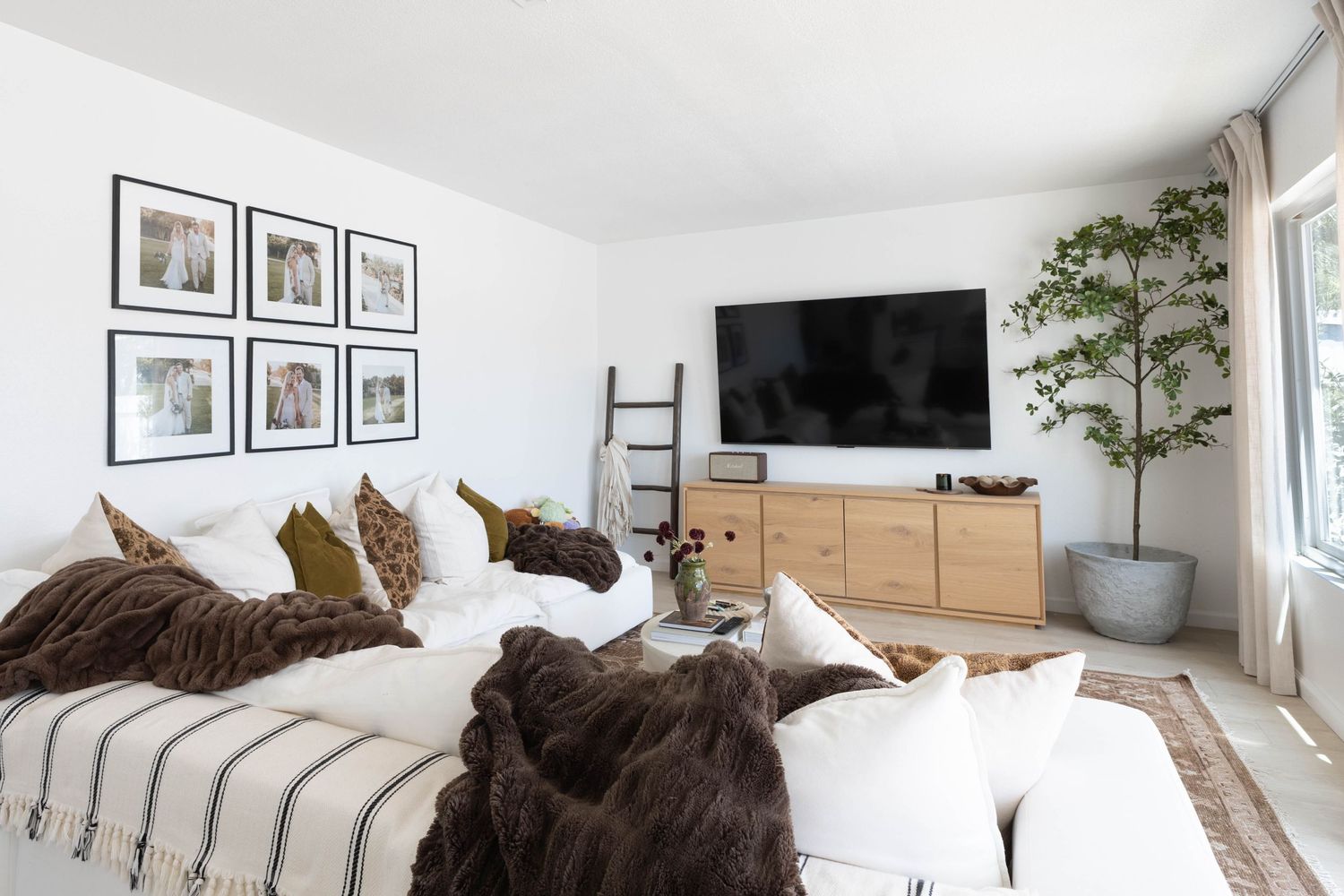 Cozy modern living room with white sectional sofa, large TV on wood console, gallery wall frames, and indoor tree decor.