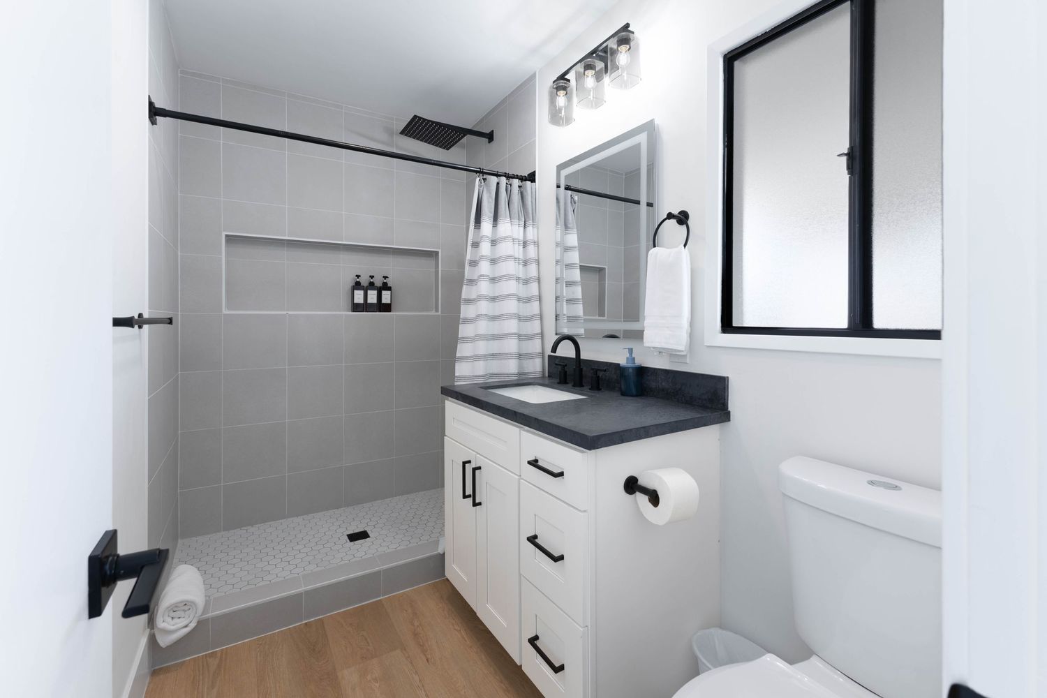 Modern bathroom with white vanity, black hardware, gray tile walk-in shower, wood floors, and dark countertop.