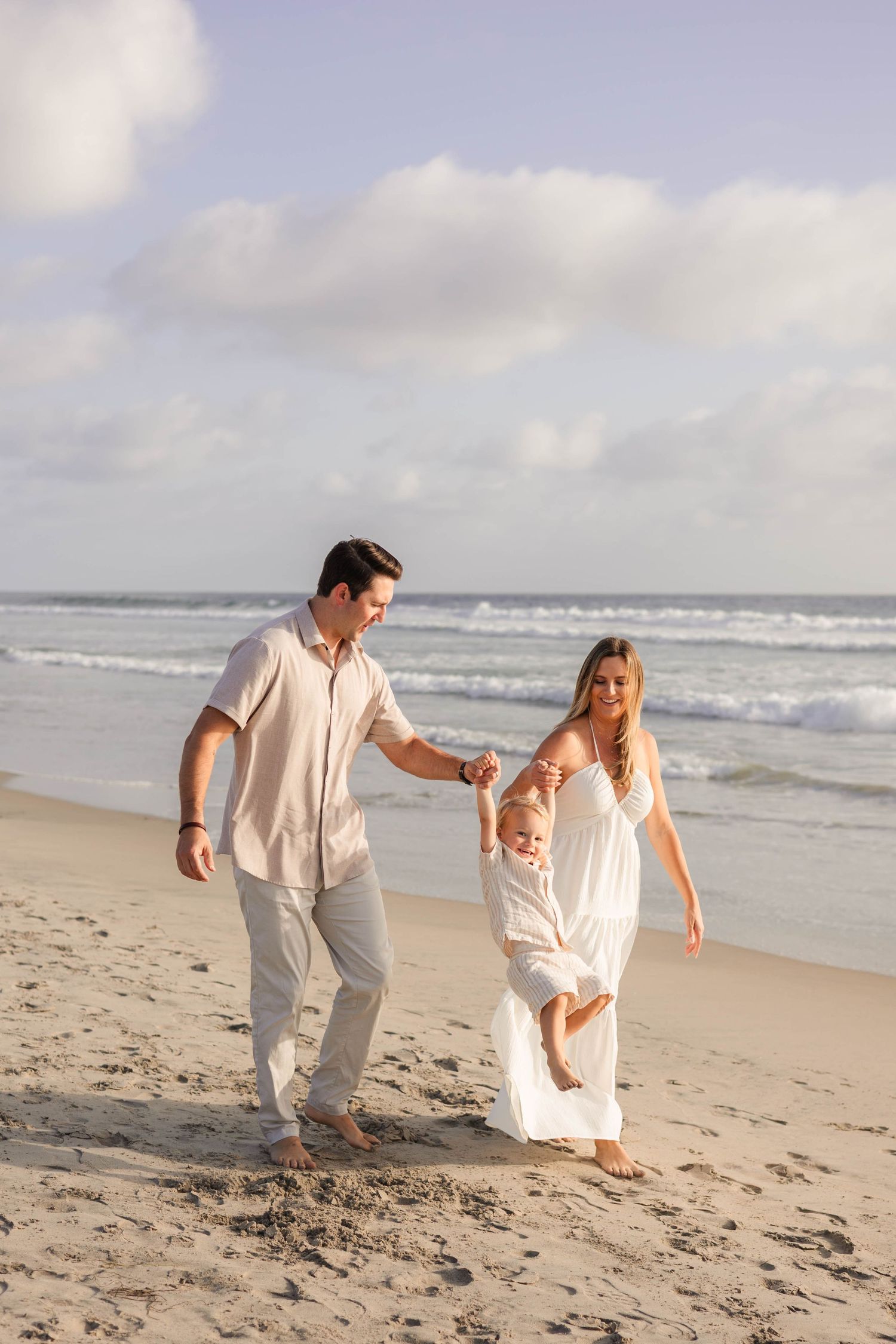 Happy family with baby enjoying a sunny beach walk, parents swinging child in white outfits by ocean waves.