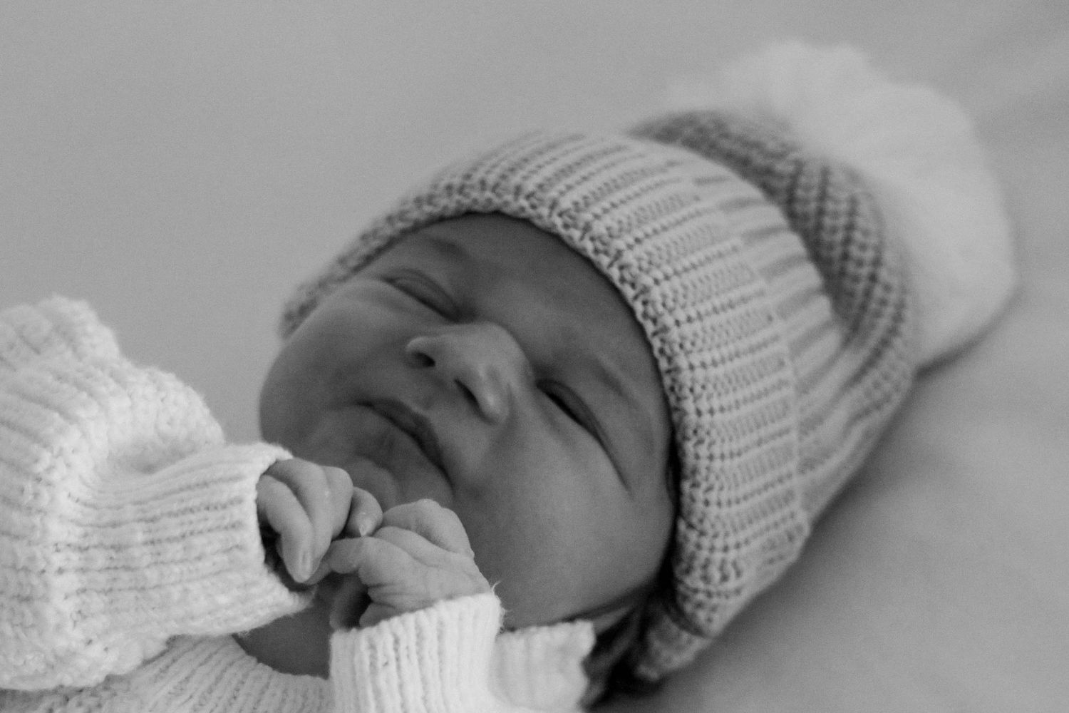 A peaceful newborn baby sleeps in a knit pompom hat in a soft black and white photo.