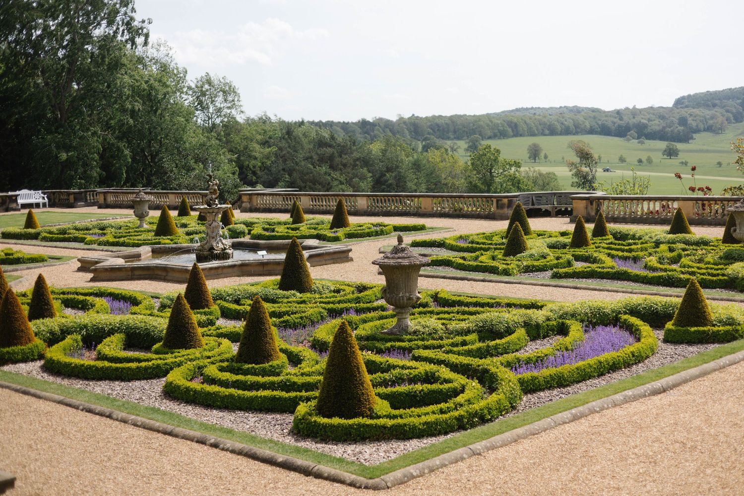 Formal English garden with geometric topiary hedges, cone-shaped yews, and ornamental urns overlooking a green valley.
