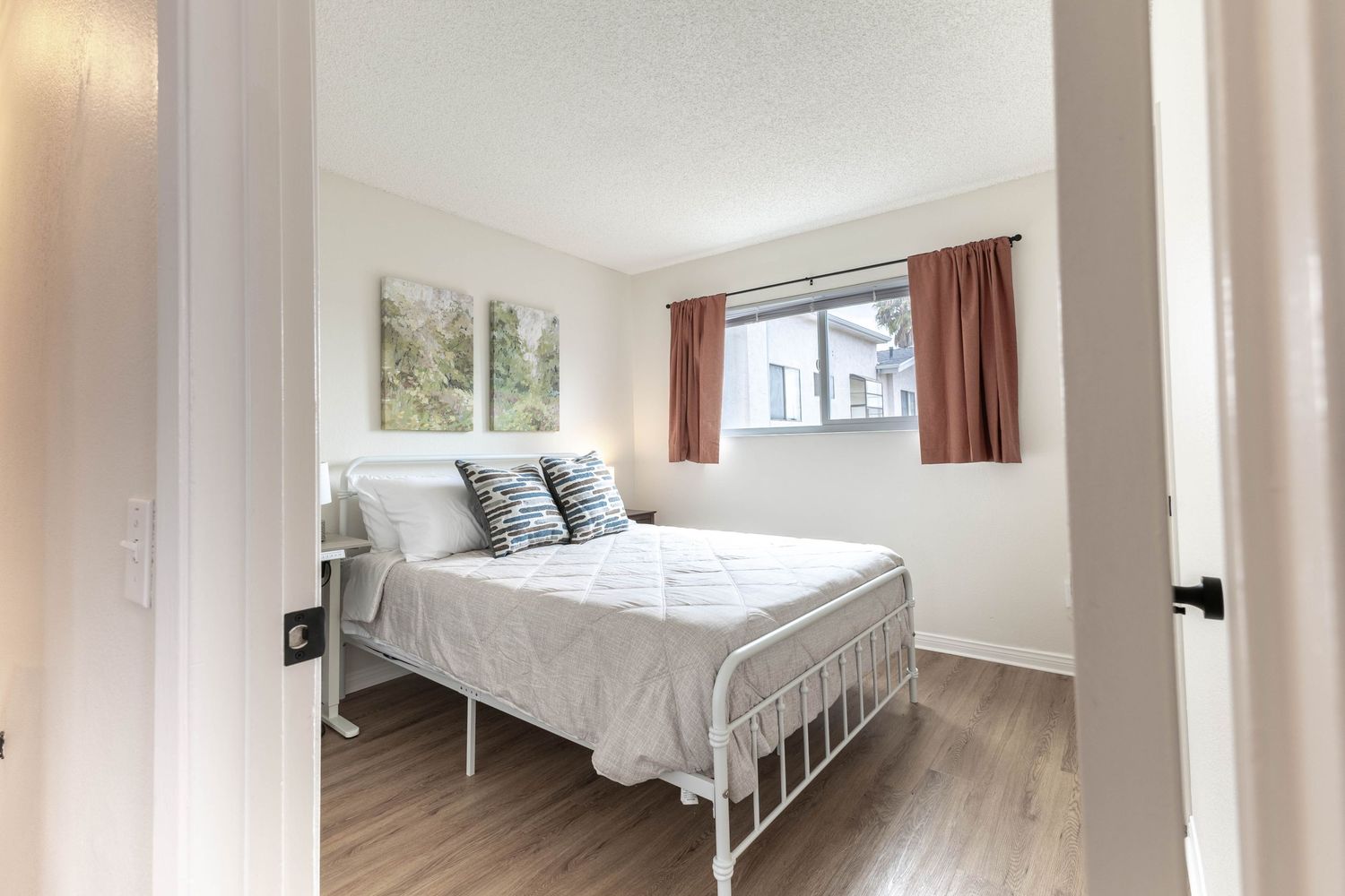 Cozy bedroom with white metal bed frame, hardwood floors, neutral walls, and warm red curtains framing a bright window.