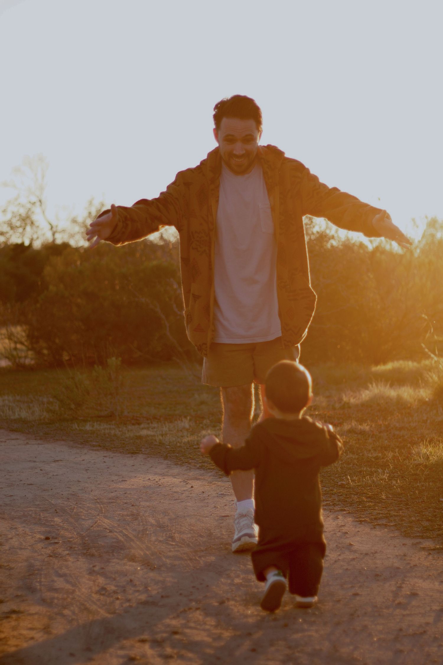 A father joyfully opens his arms as his young child runs toward him on a sunlit dirt path.