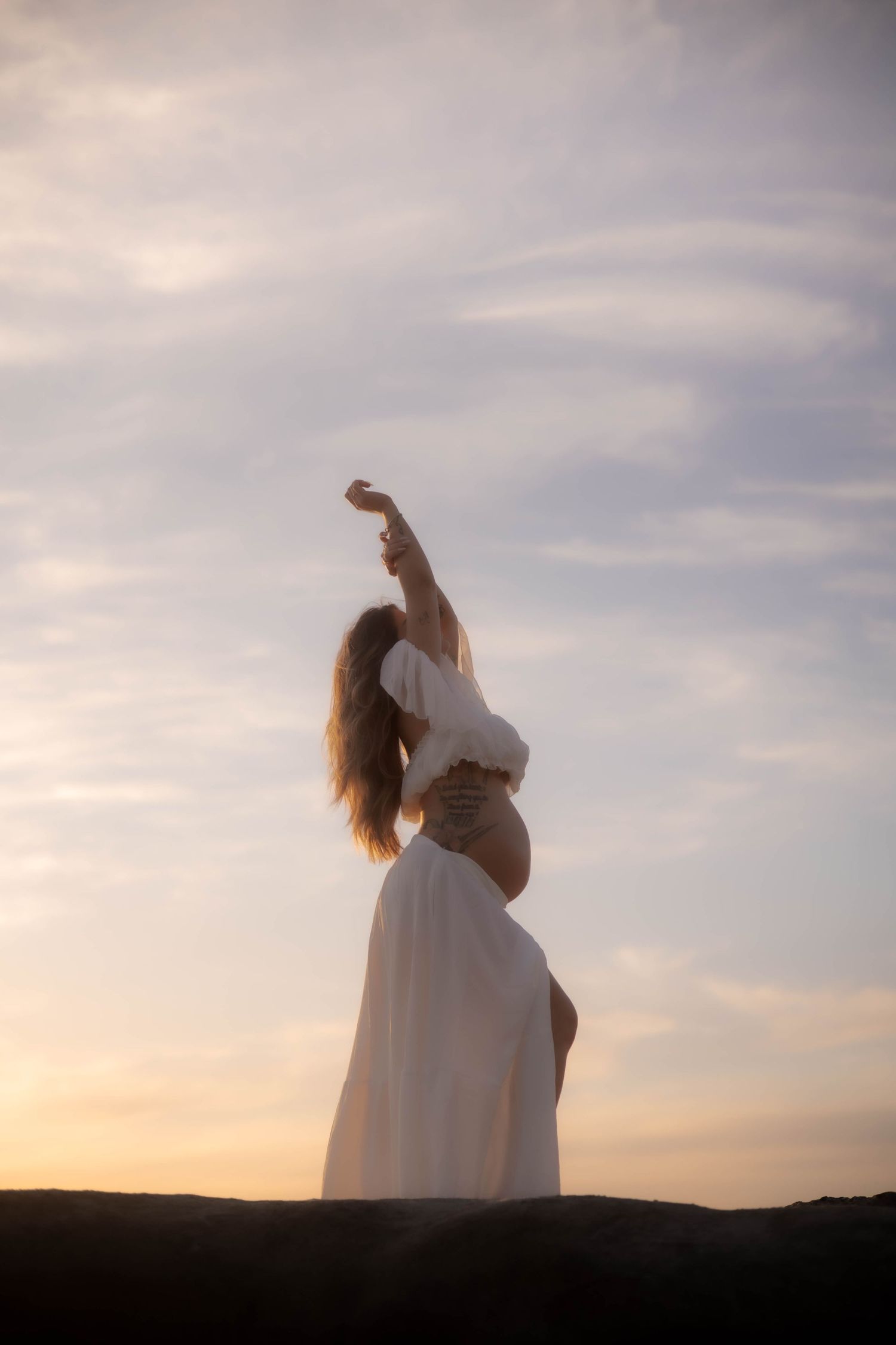 Silhouette of pregnant woman in white dress with arm raised against dramatic sunset sky.