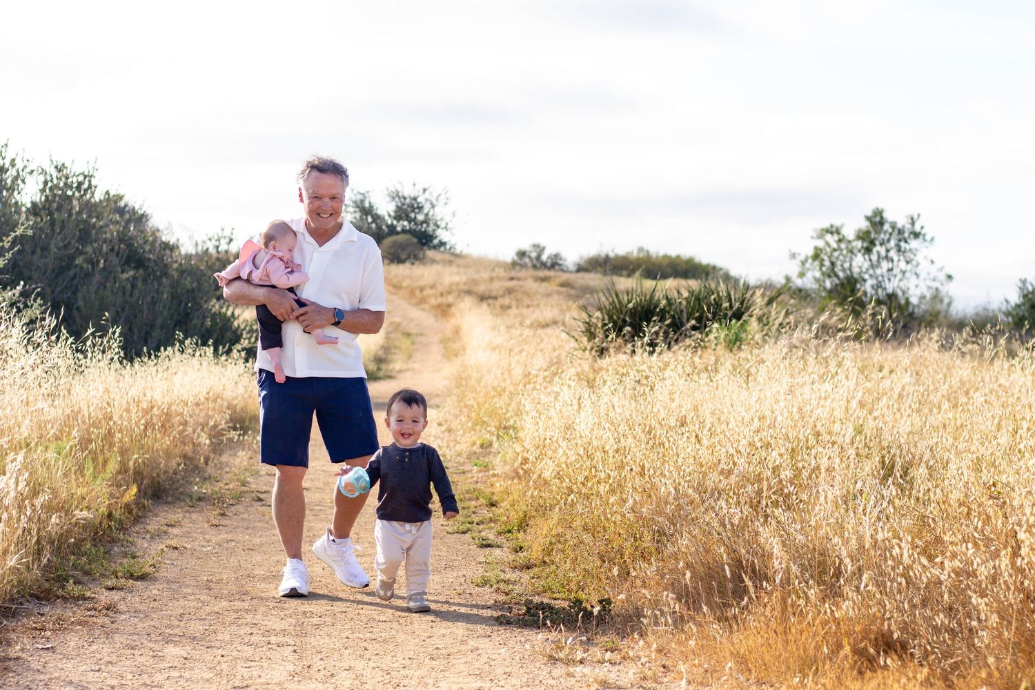 Father holding infant while walking with toddler on sunny golden grass trail outdoors.