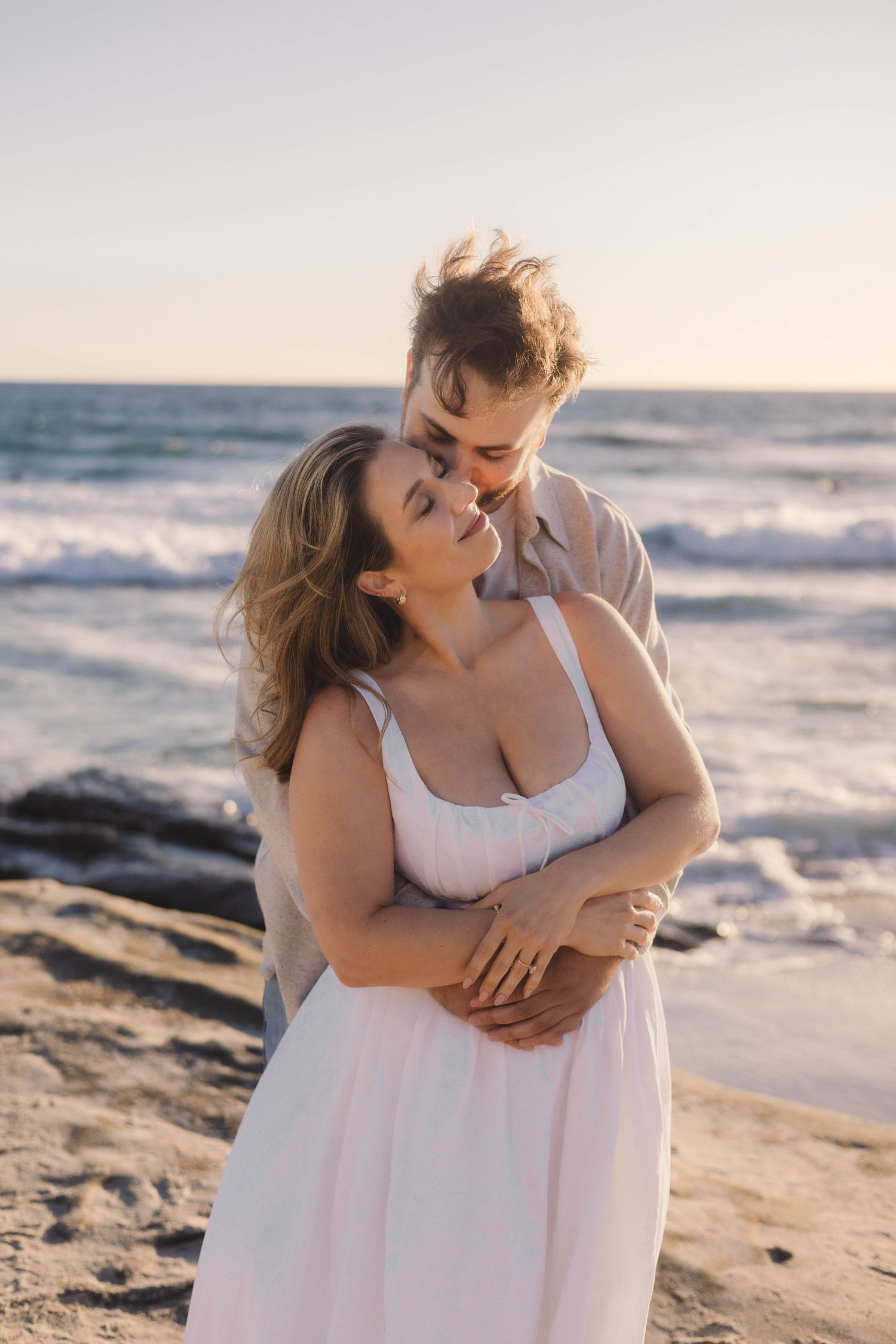 Couple embracing romantically on a sandy beach at sunset with ocean waves in the background.