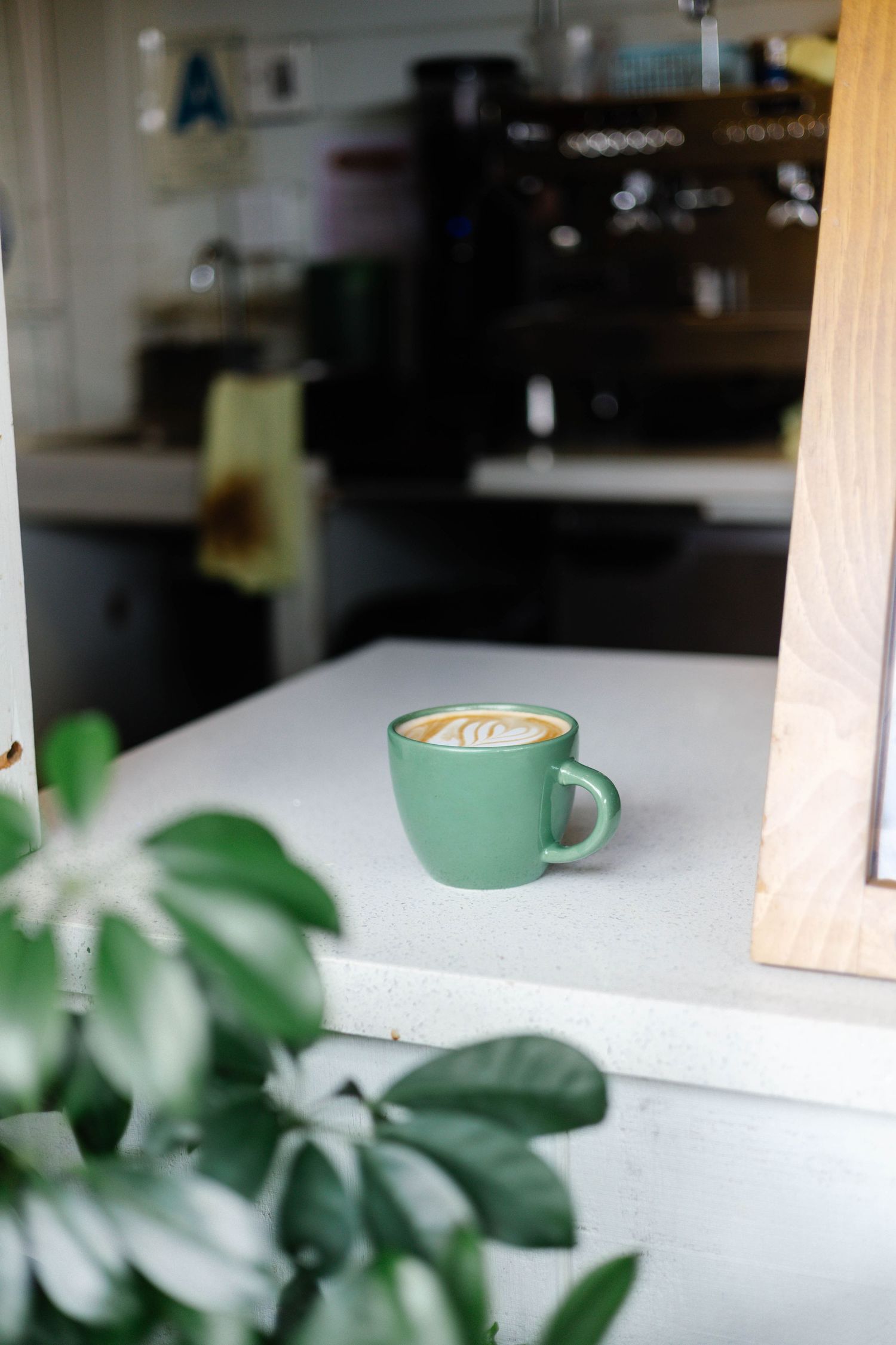 A green ceramic latte cup with latte art sits on a white counter inside a cozy cafe.