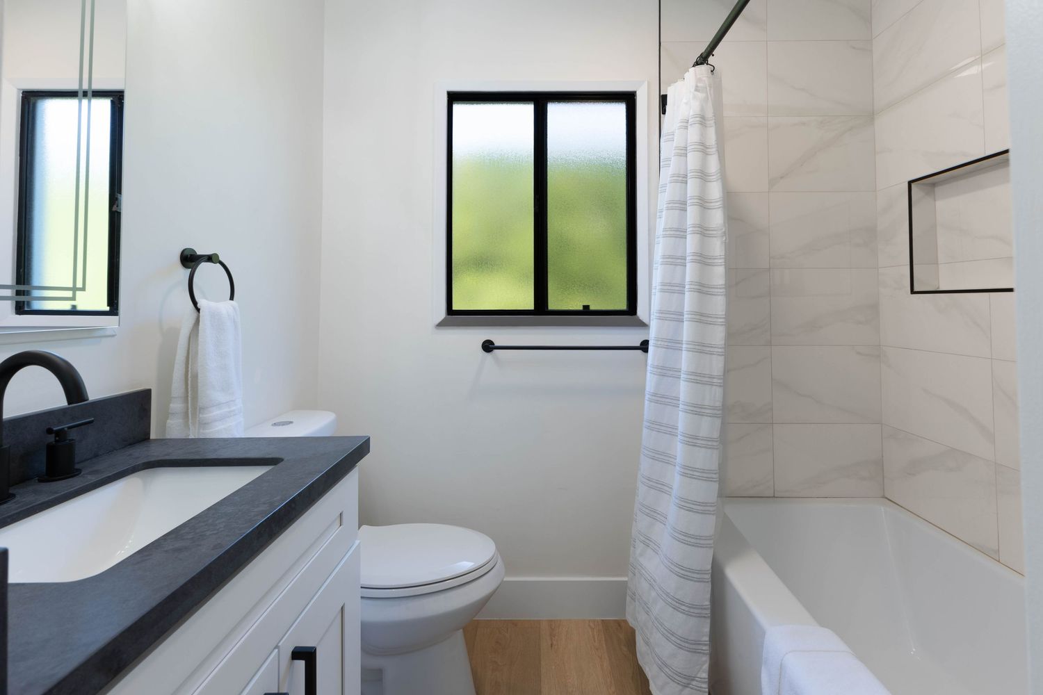 Modern bathroom with black fixtures, white vanity, gray countertop, tiled shower-tub combo, and natural light from window.