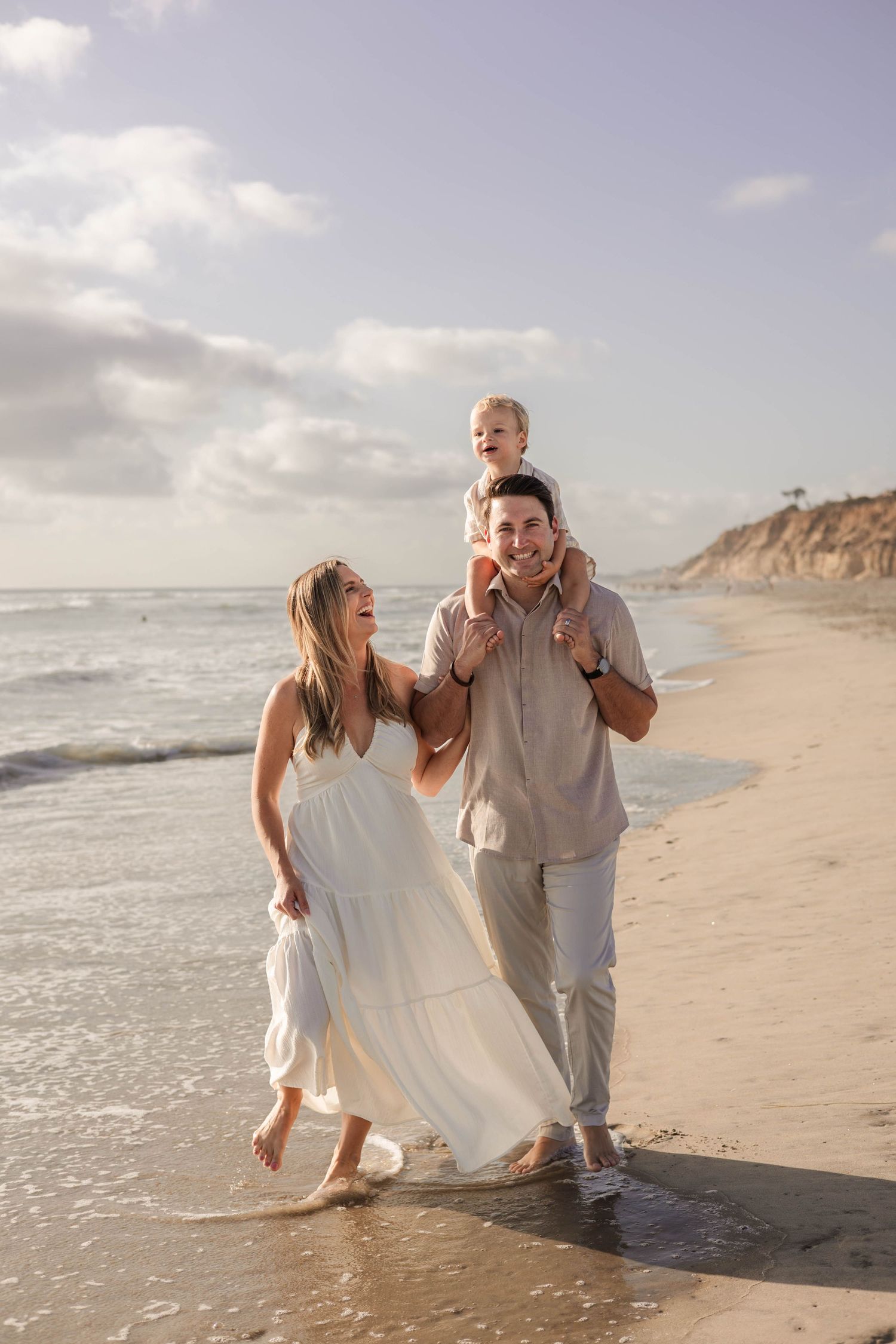 Family of three walking on beach, parents smiling as toddler sits on dad's shoulders, wearing neutral tones.