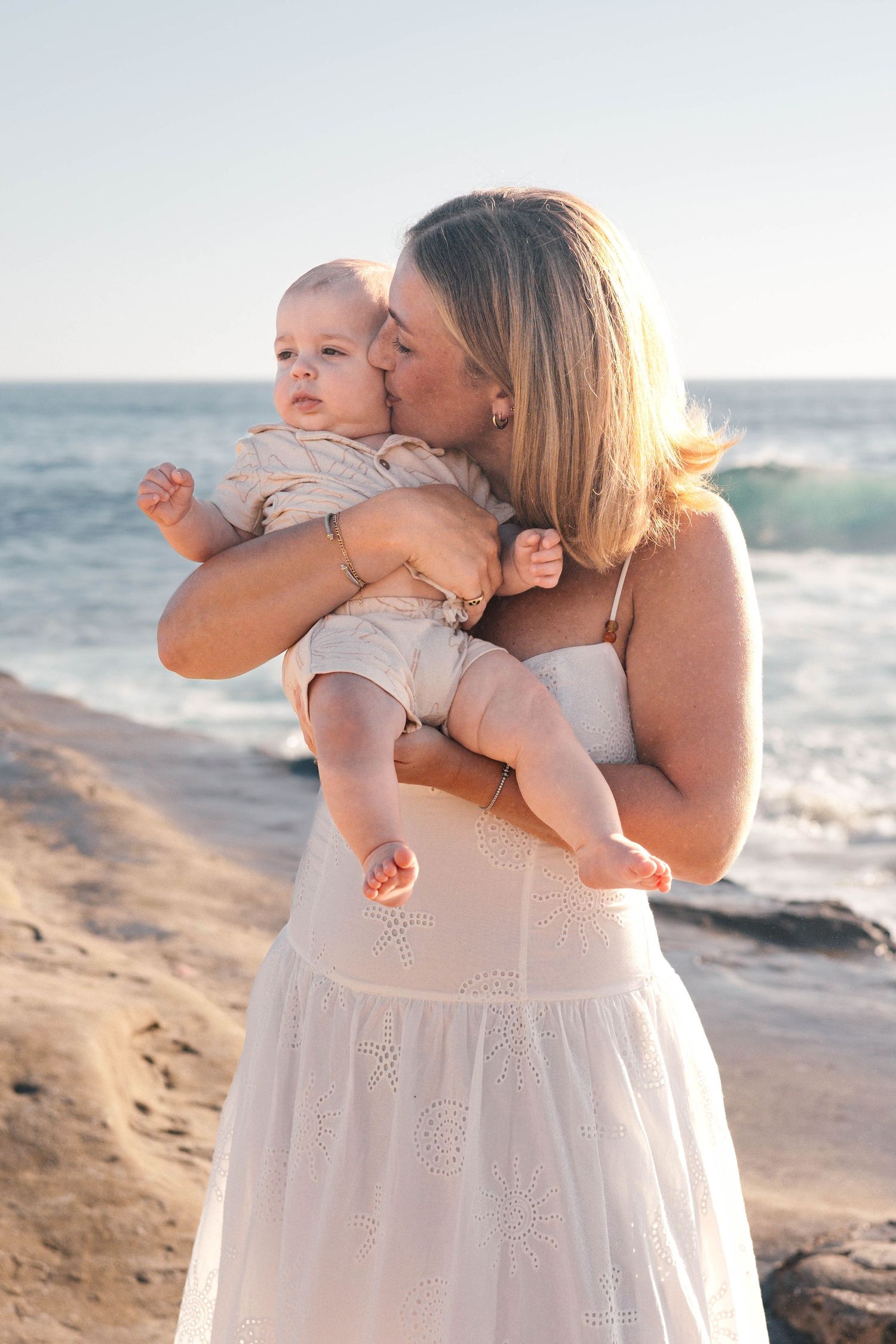 A mother in a white dress kisses her baby on the cheek while standing on a rocky beach with ocean waves behind.