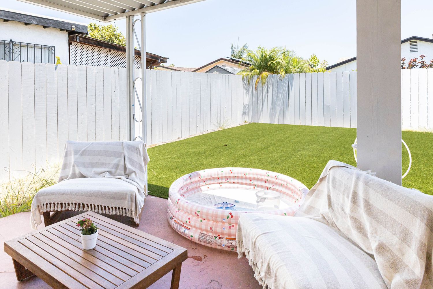 Cozy backyard patio with white fence, lush green lawn, inflatable pool, outdoor sofa, and wooden coffee table.