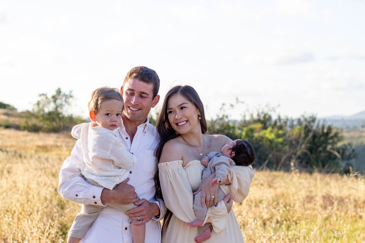 Happy family of four poses in golden field at sunset, parents holding toddler and newborn baby.
