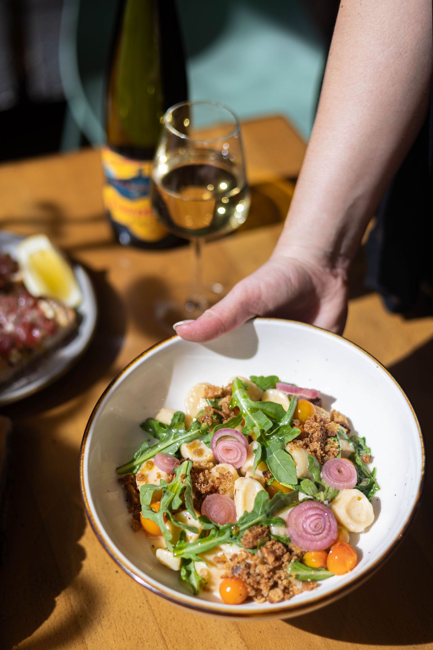 A hand placing a fresh salad bowl with arugula, vegetables, and grains beside a glass of white wine on a sunny table.