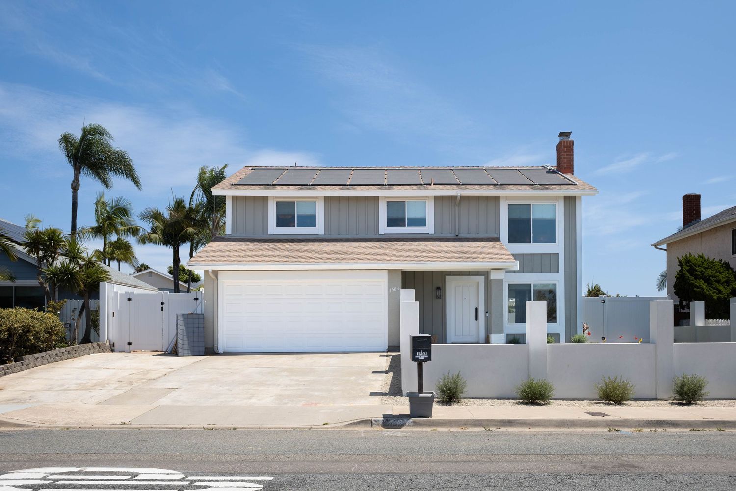 Two-story coastal home with white garage door, solar panels, palm tree, and white fence on a sunny day.