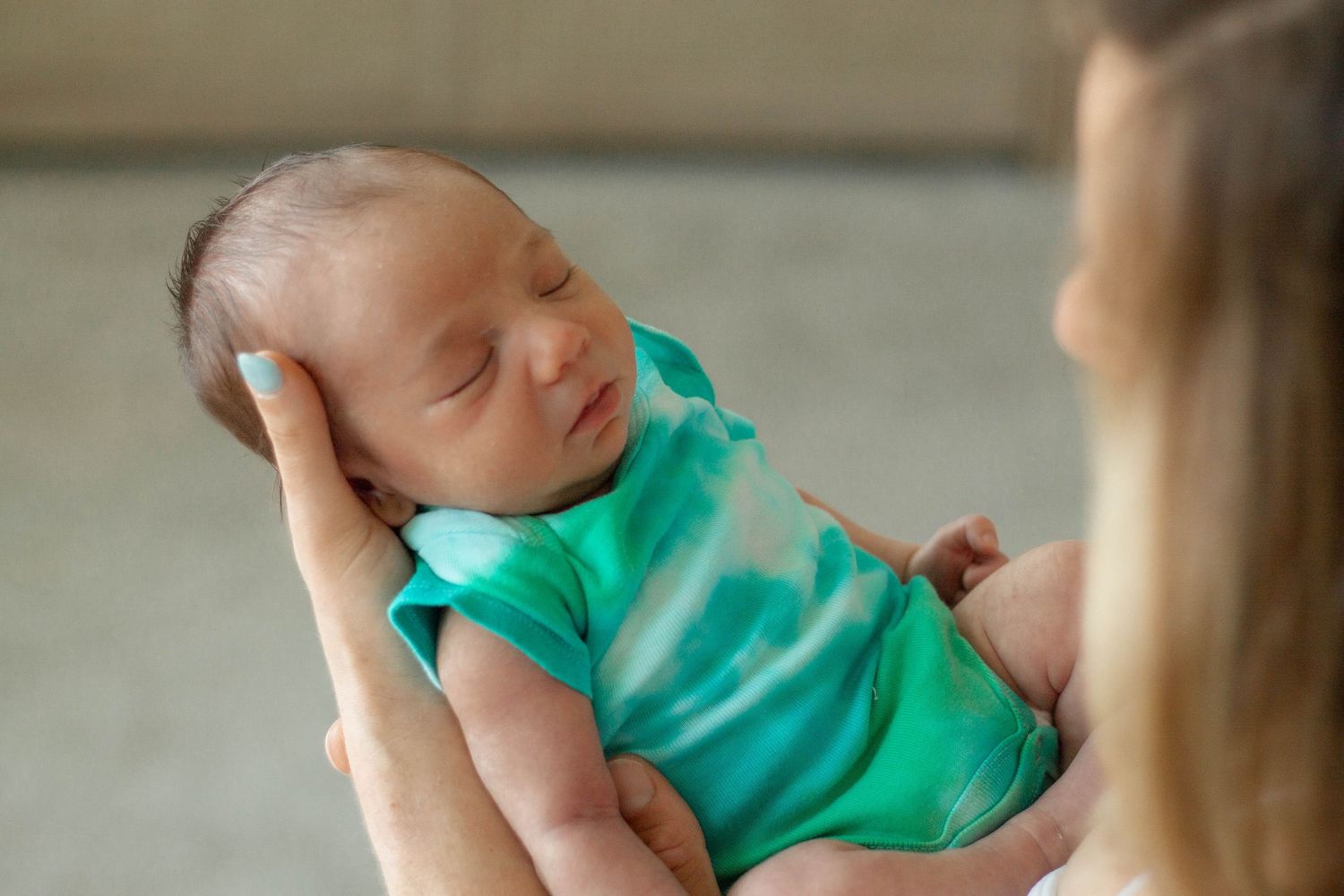 Mother tenderly holds sleeping newborn baby dressed in teal onesie, cradling infant's head gently in her hands.